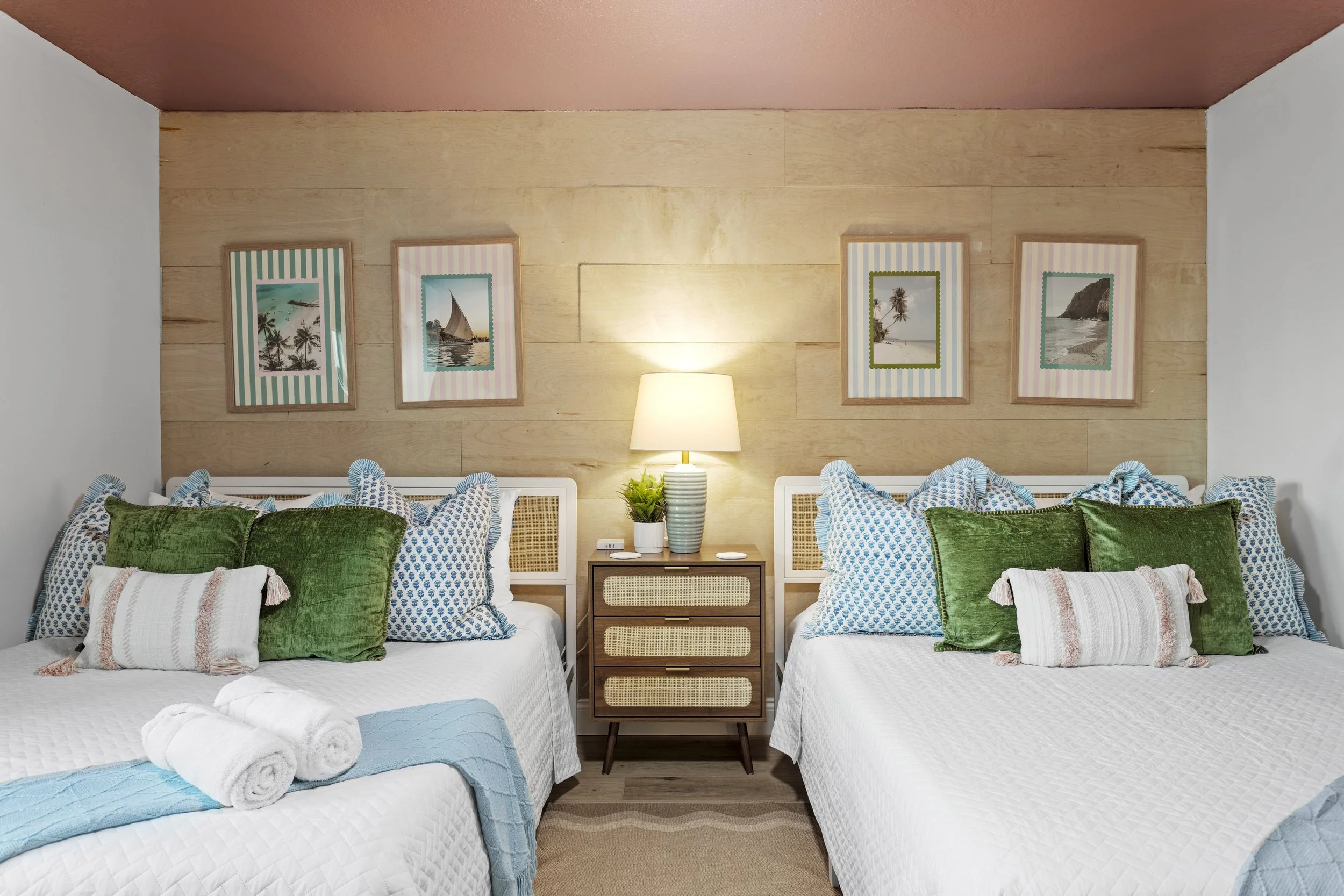 A bedroom with two beds, each with white quilted bedspreads, multiple green and blue patterned pillows, and rolled white towels on one bed. A wooden nightstand with drawers and a lamp with a striped shade is between the beds. The back wall features framed pictures of beach scenes and has a wood panel decor. The ceiling is pink, and the room has a cozy, tropical vibe.
