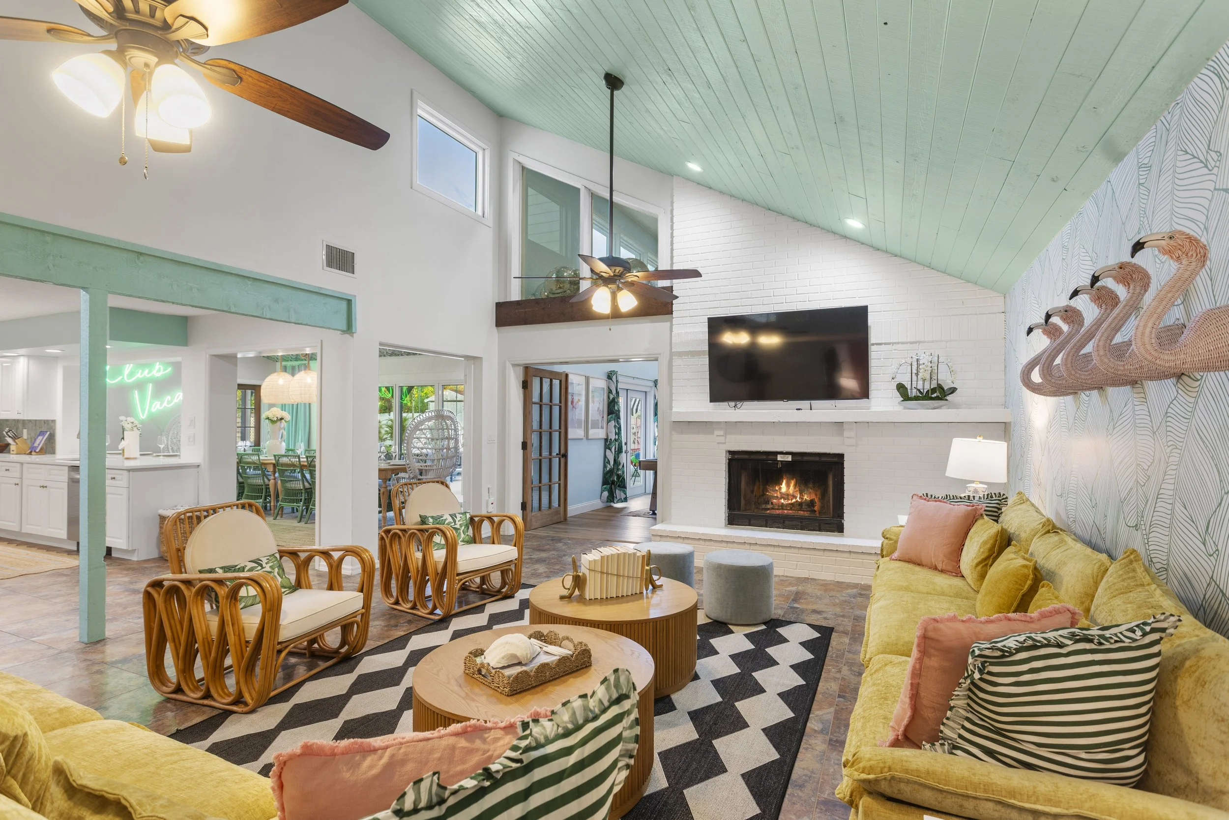 Living room with yellow sofas, a white brick fireplace, wall-mounted TV, wooden coffee tables, decorative flamingo wall art, ceiling fans, green and white wallpaper, and large windows.