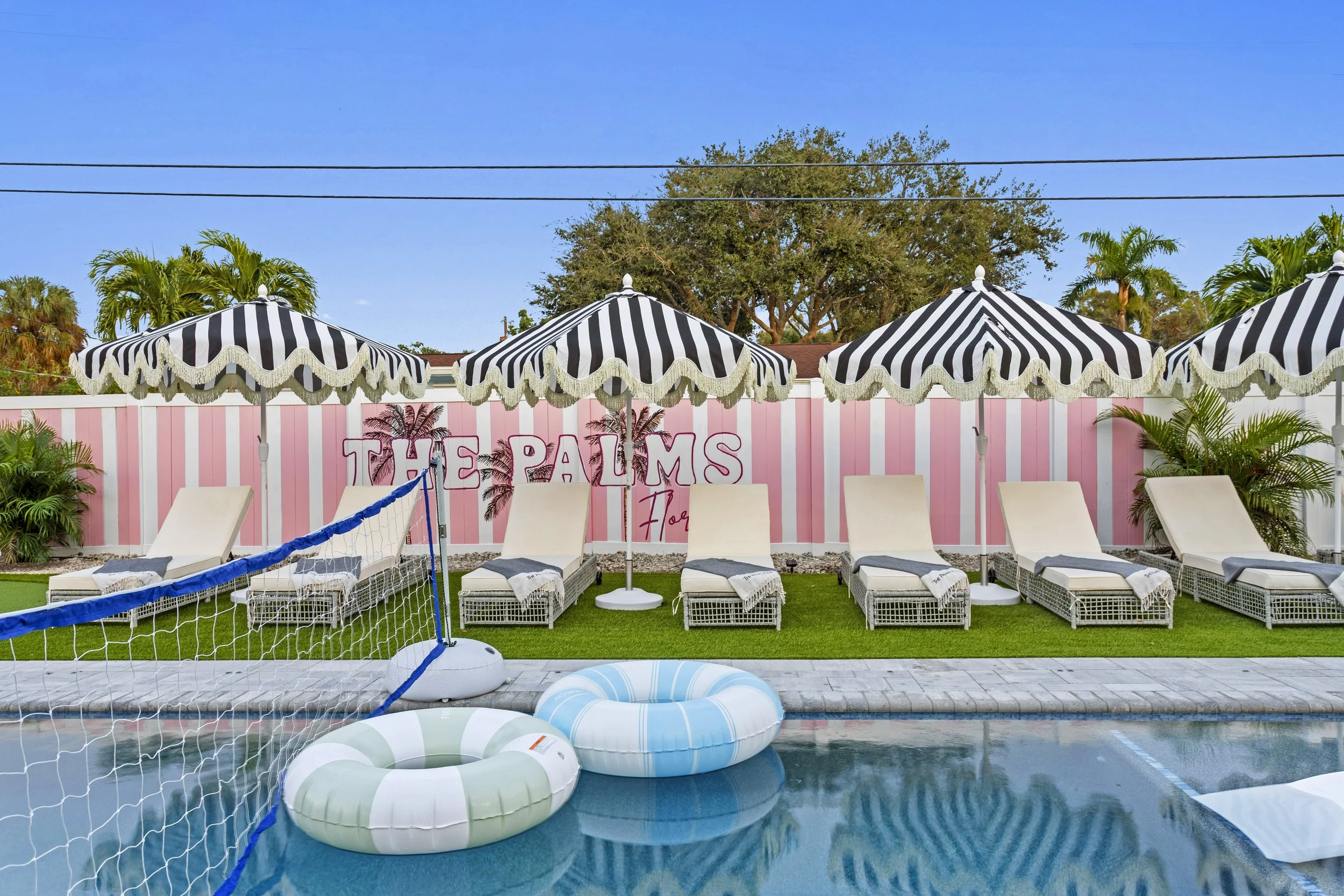 Poolside area with lounge chairs, umbrellas, and pink striped wall with sign that says 'The Palms' in pink neon lights