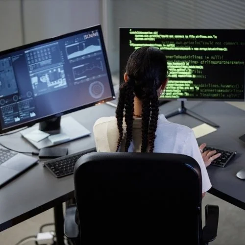 Person with braided hair working at a computer with multiple monitors displaying graphs and green code on the screen