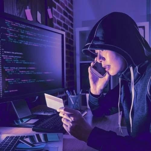 A hacker in a hoodie talking on a cell phone in a dimly lit room with computer coding on the screen.