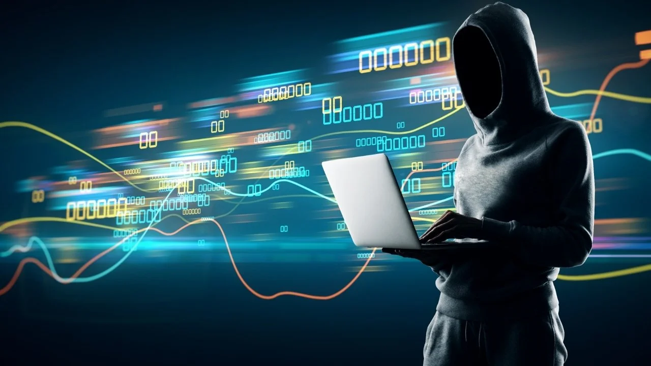 A hooded figure using a laptop with digital financial or stock market graphs and data in the background.