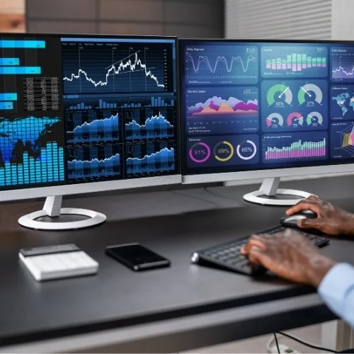 Three computer monitors displaying stock market graphs, charts, and data visualizations. A person's hands are on a keyboard and mouse in front of the monitors. There is a notebook and a phone on the desk.
