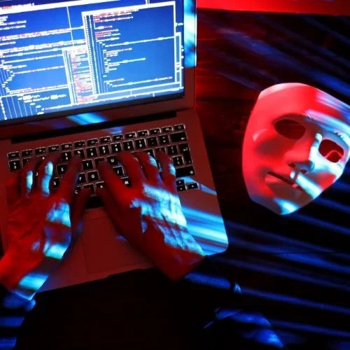 A mystery person typing on a laptop with blue coding screens and lines of code, with a dark background and red lighting. Beside them is a white mask representing their hidden identity, eluding to a hacker.