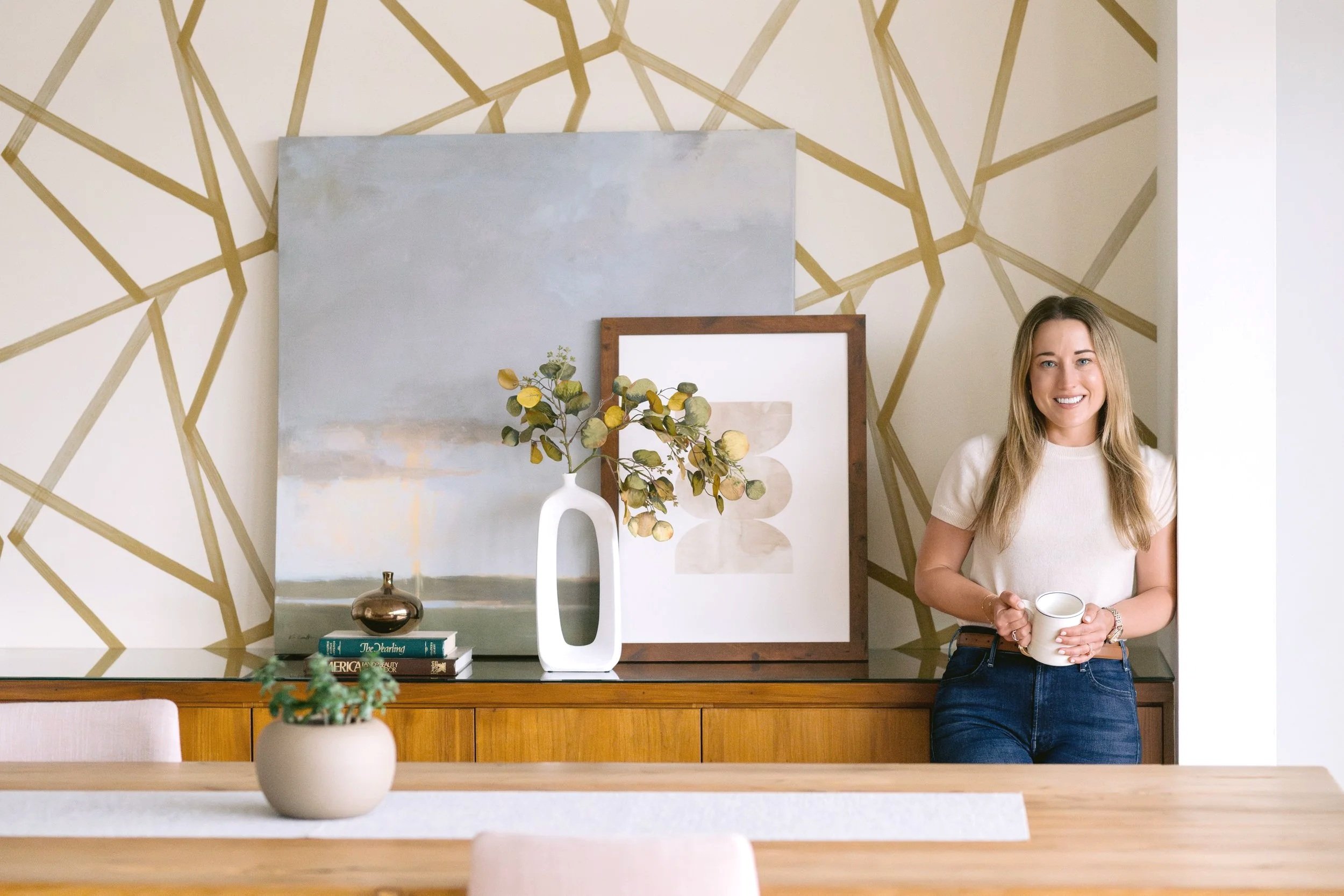 A woman with long blonde hair wearing a white shirt holding a white mug, standing in a modern room with abstract wall art and decorative items on the wooden table behind her.