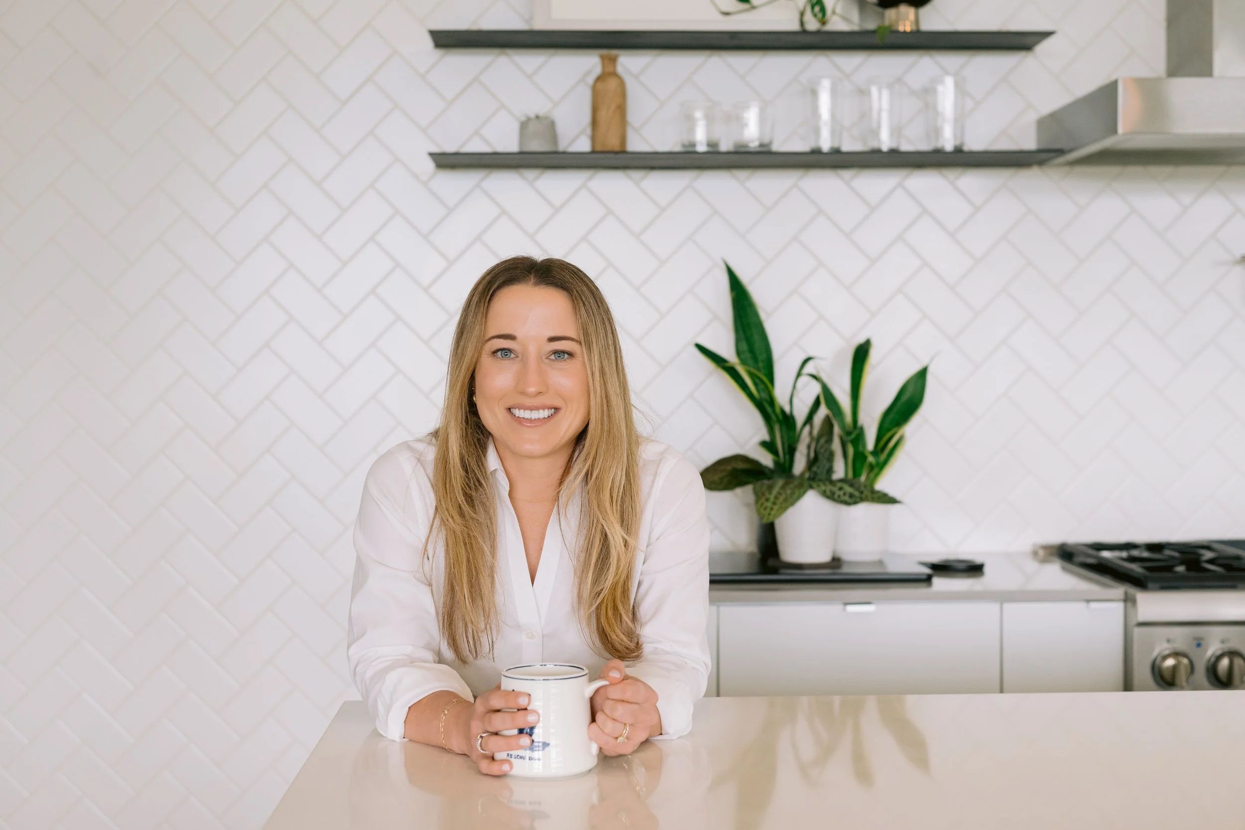 A woman with blonde hair smiling while holding a white mug in a modern kitchen with white tiled walls, green plant, and black shelves.