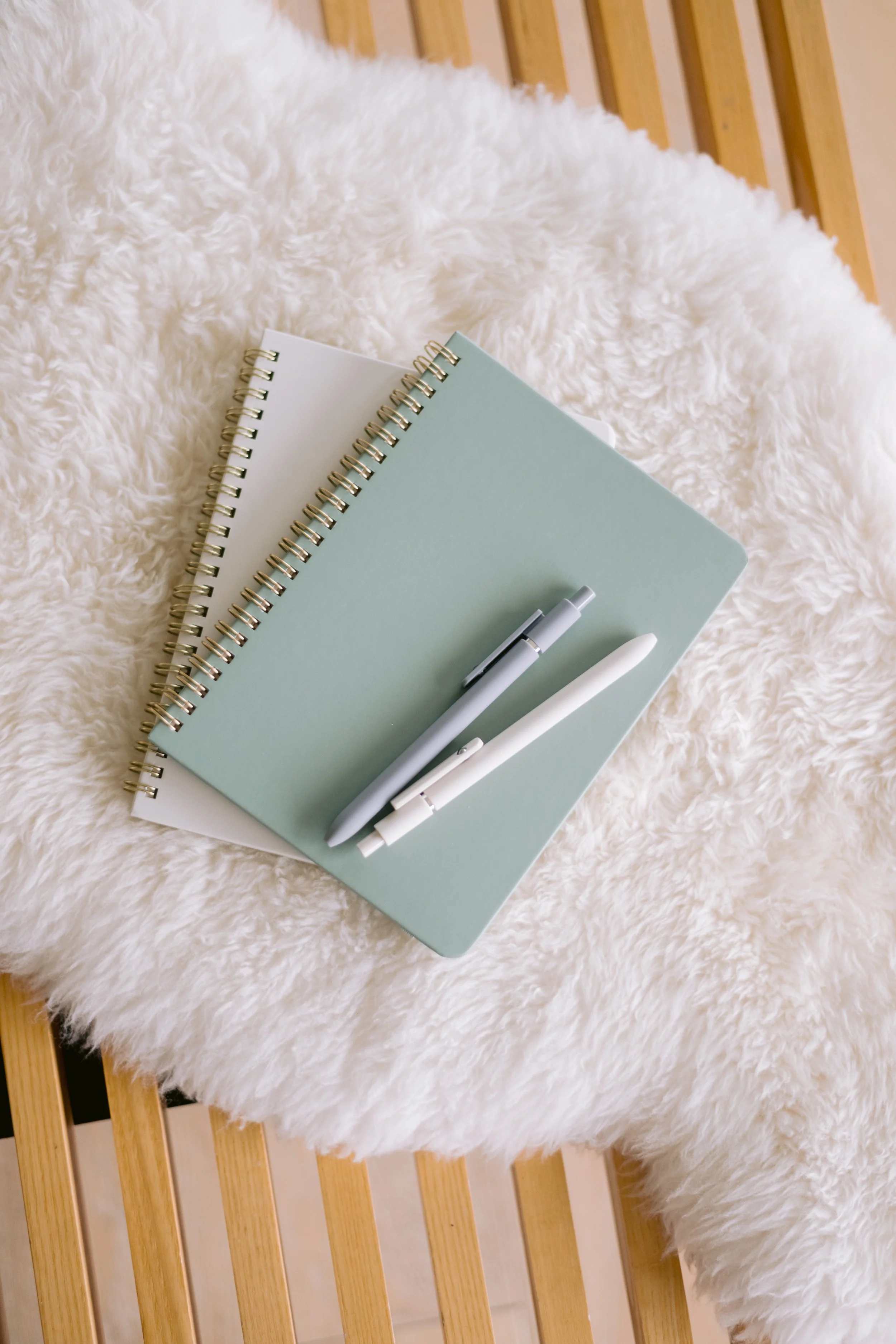 Two spiral-bound notebooks, one white and one pastel blue, with a white pen and a white marker resting on top, placed on a soft, fluffy white surface with a wooden frame in the background.