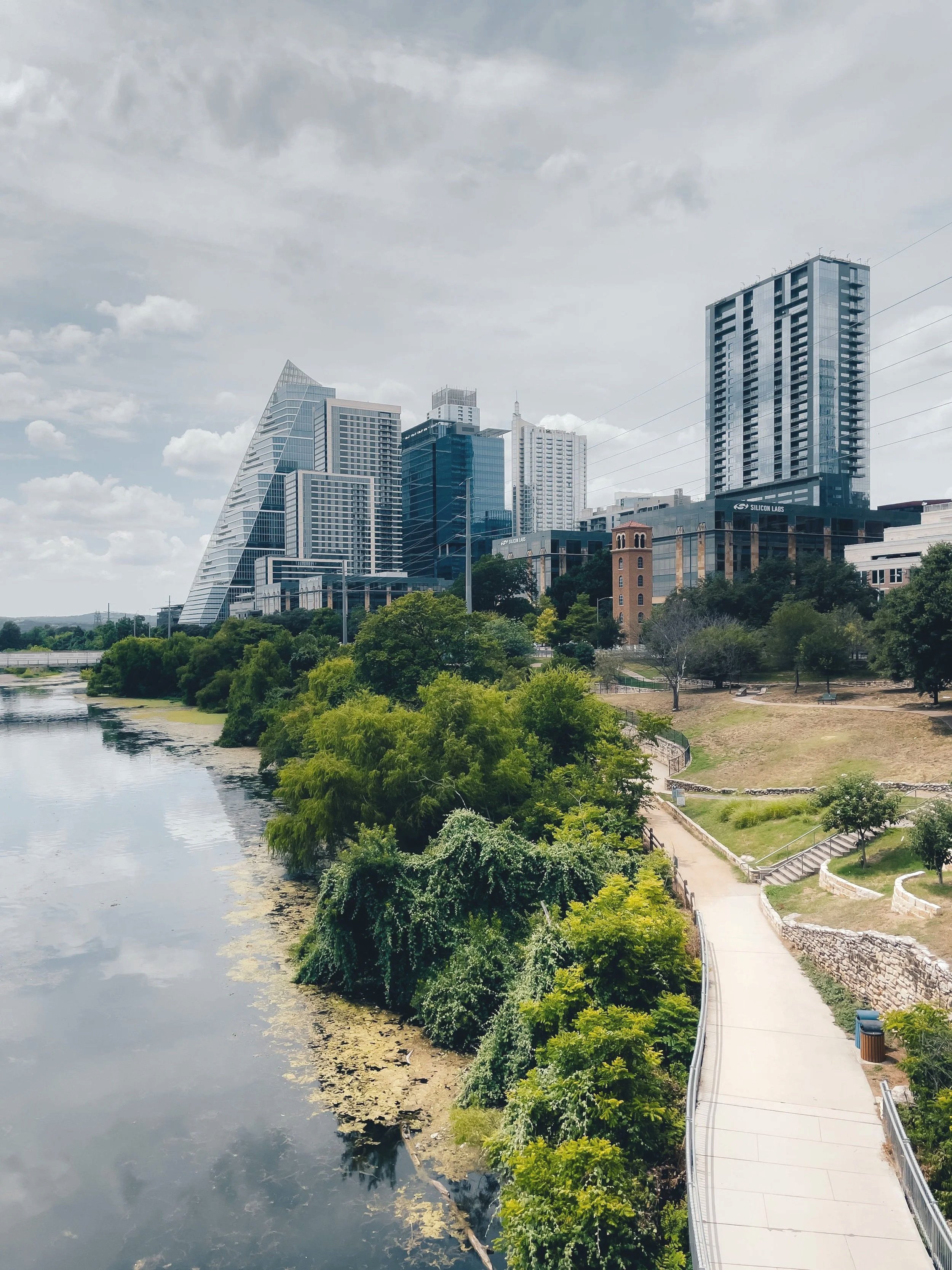View of a city skyline with modern high-rise buildings near a river, green trees, and a walking path along the riverbank.