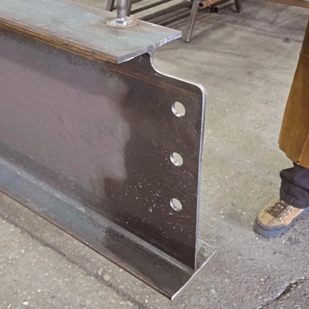 Close-up of a metal flat bar with three holes, sitting on a workbench in a workshop.