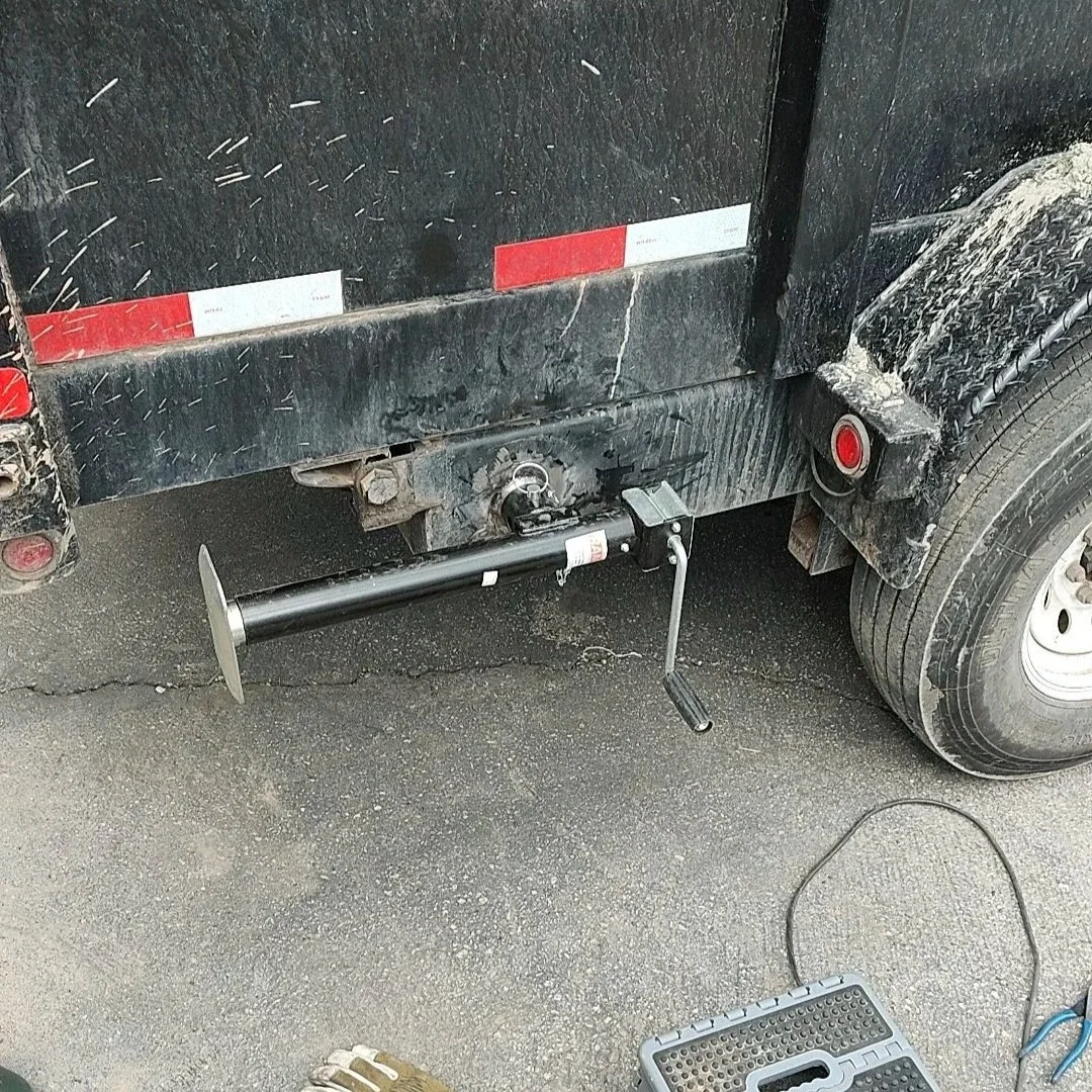 Close-up of a trailer hitch being installed on a black trailer, with welding equipment and gloves on the ground nearby.