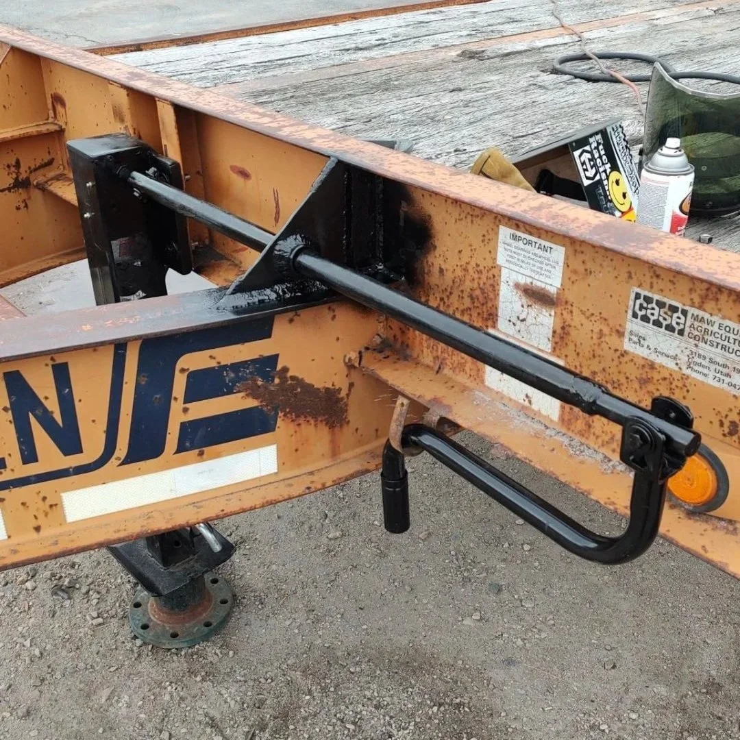 A construction site featuring a trailer with rust and black paint, with a large pipe elevated in the background, and construction tools and equipment scattered around, including a workman in an orange shirt and grey hat.
