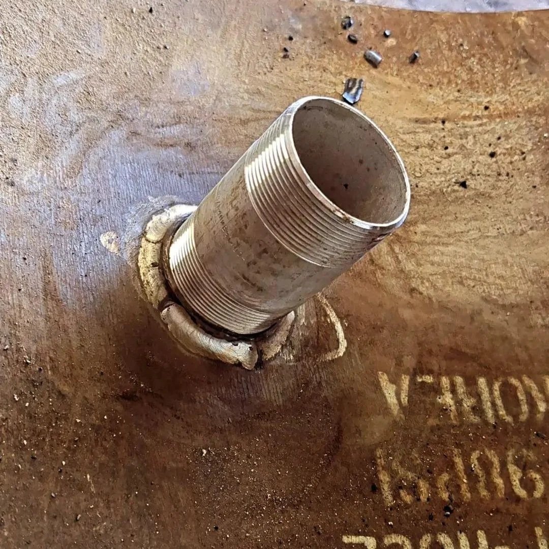 A metal pipe protruding from a large metal surface, with visible threading on the pipe and some surrounding residue, along with dirt and debris nearby.