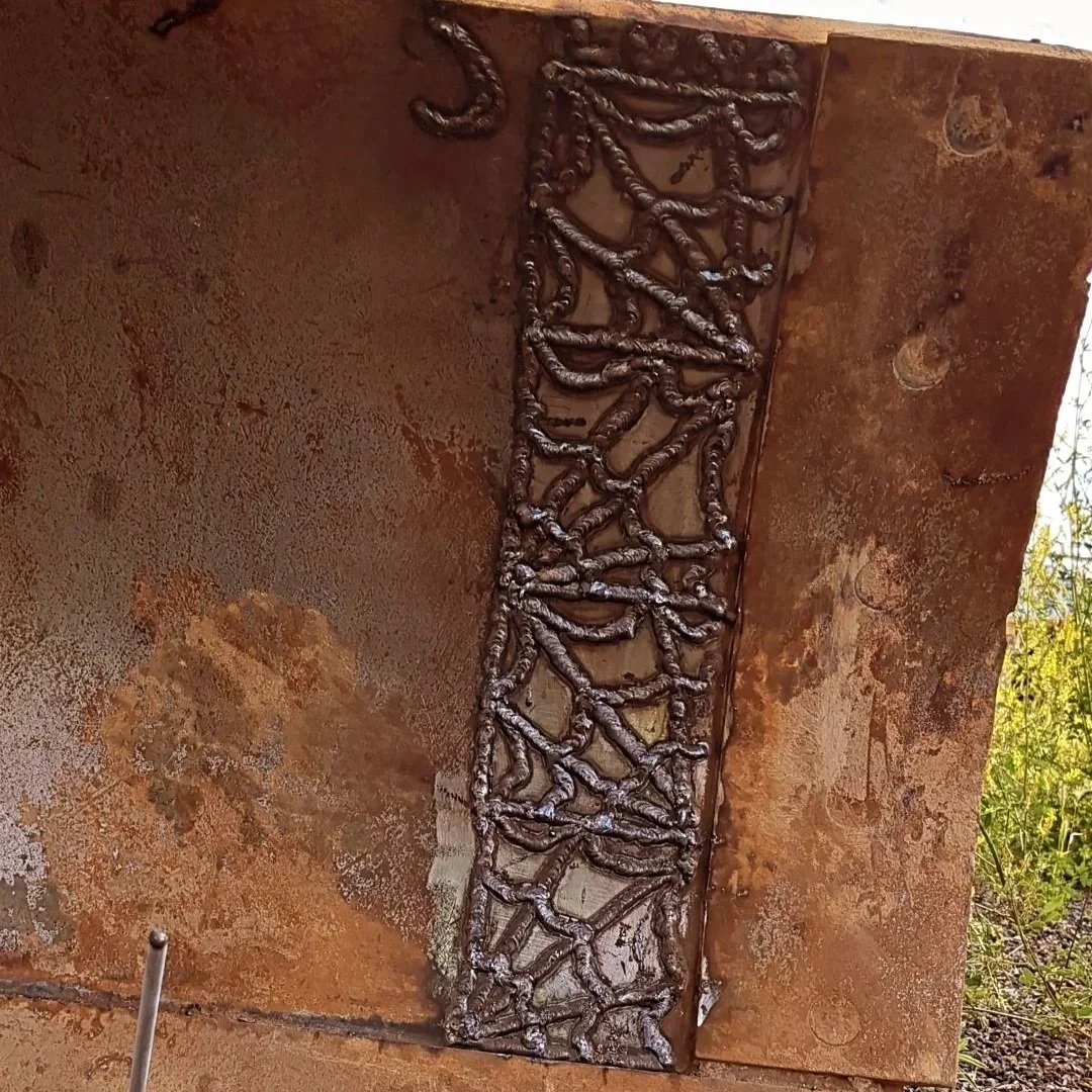 Close-up view of a metal plate with intricate weld patterns, part of a construction or machinery project, with tools and gravel on the ground.