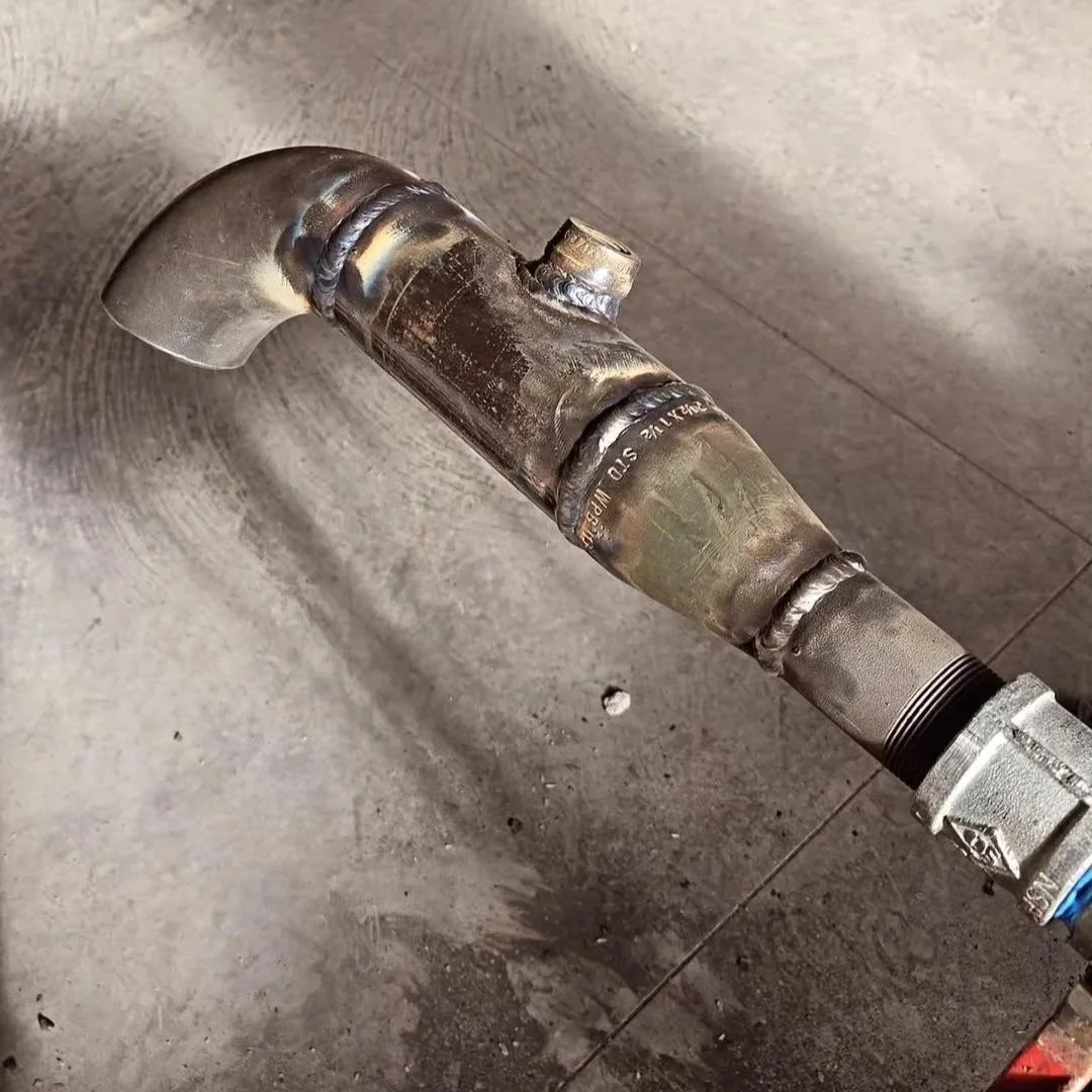 A metallic pipe with welding marks, resting on a concrete floor in a workshop or construction site.