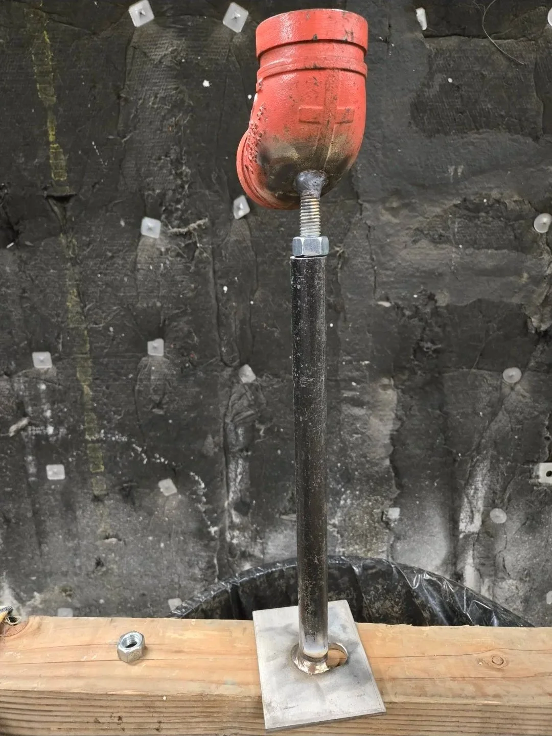A makeshift valve or weather vane constructed from a red plastic or metal object mounted on a metal pole, attached to a wooden base, against a dark, textured background.