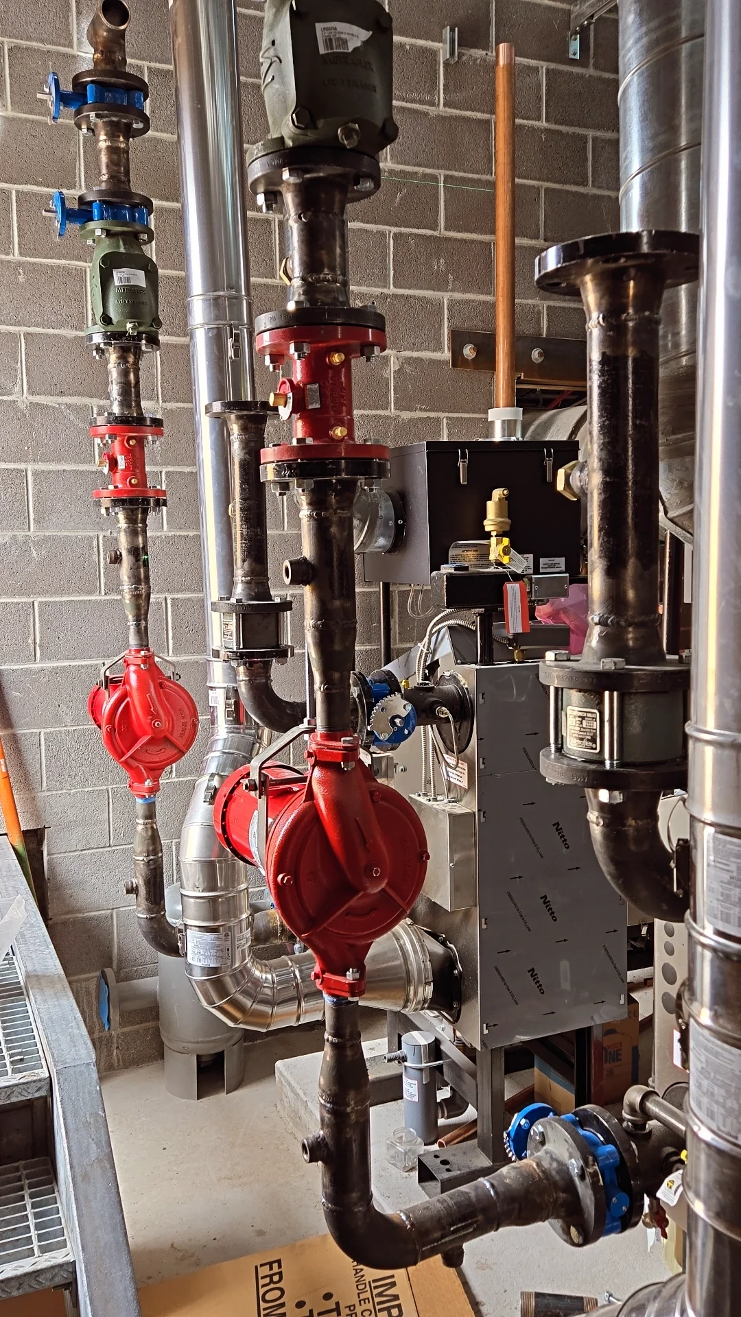 Industrial plumbing system with various pipes, valves, and machinery against a brick wall.