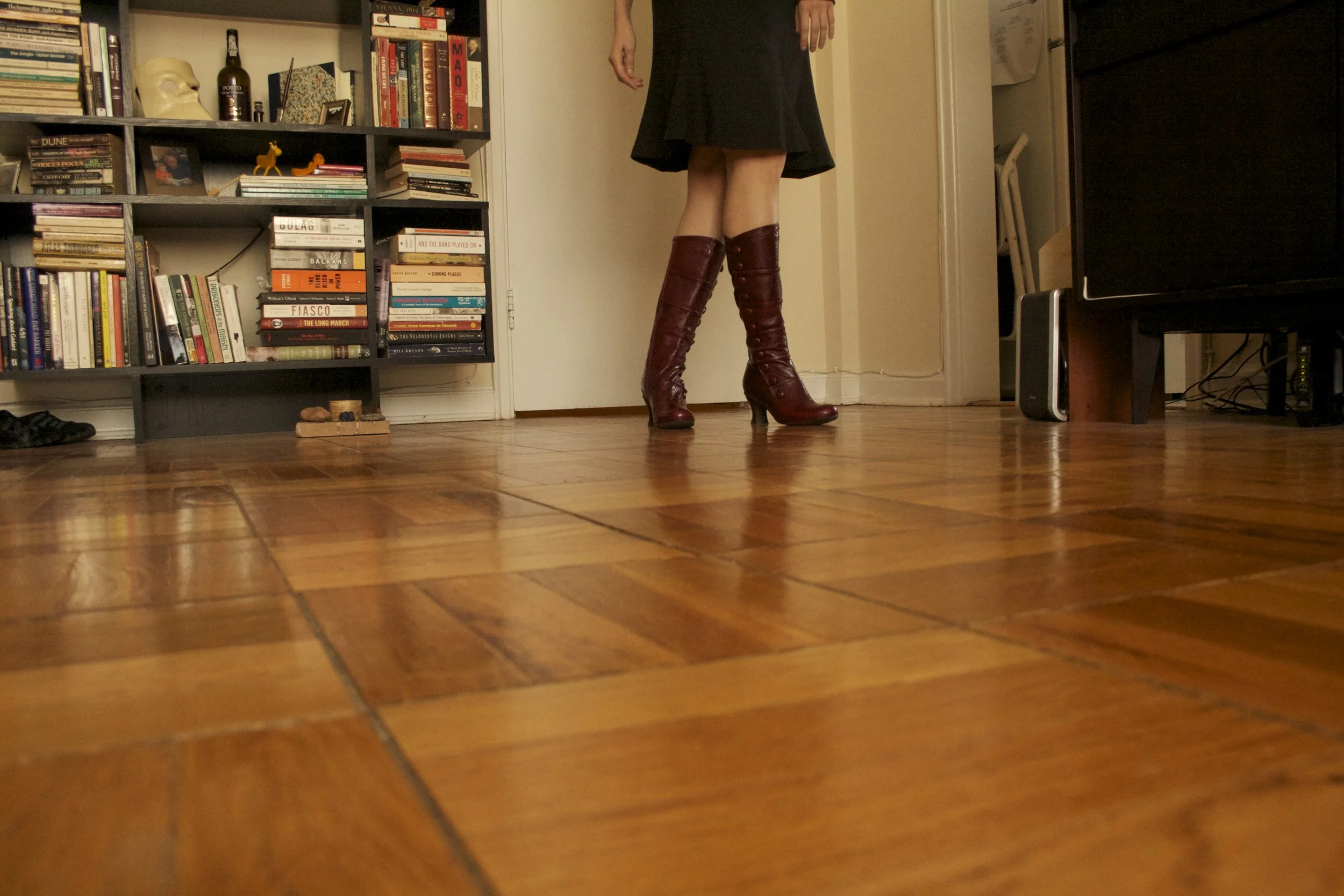 A person standing in a room wearing a black dress and knee-high burgundy boots. The room has a bookshelf filled with books and decorative items, a hardwood floor, and a door.