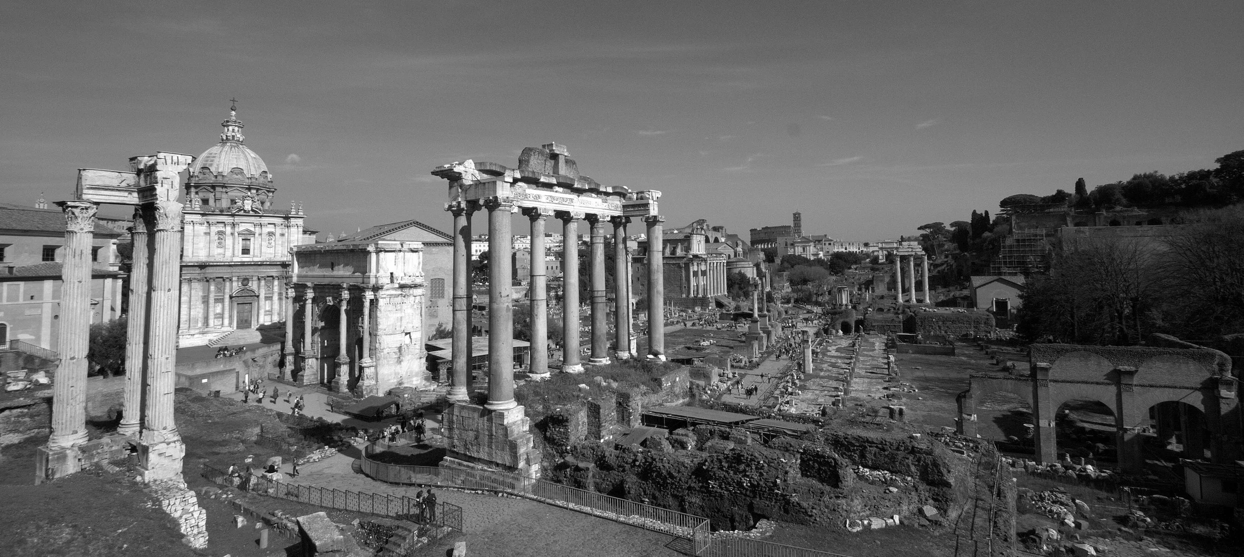 Ancient ruins of Roman temples with tall columns in a cityscape, some buildings with domes and trees visible in the background, black and white photo.