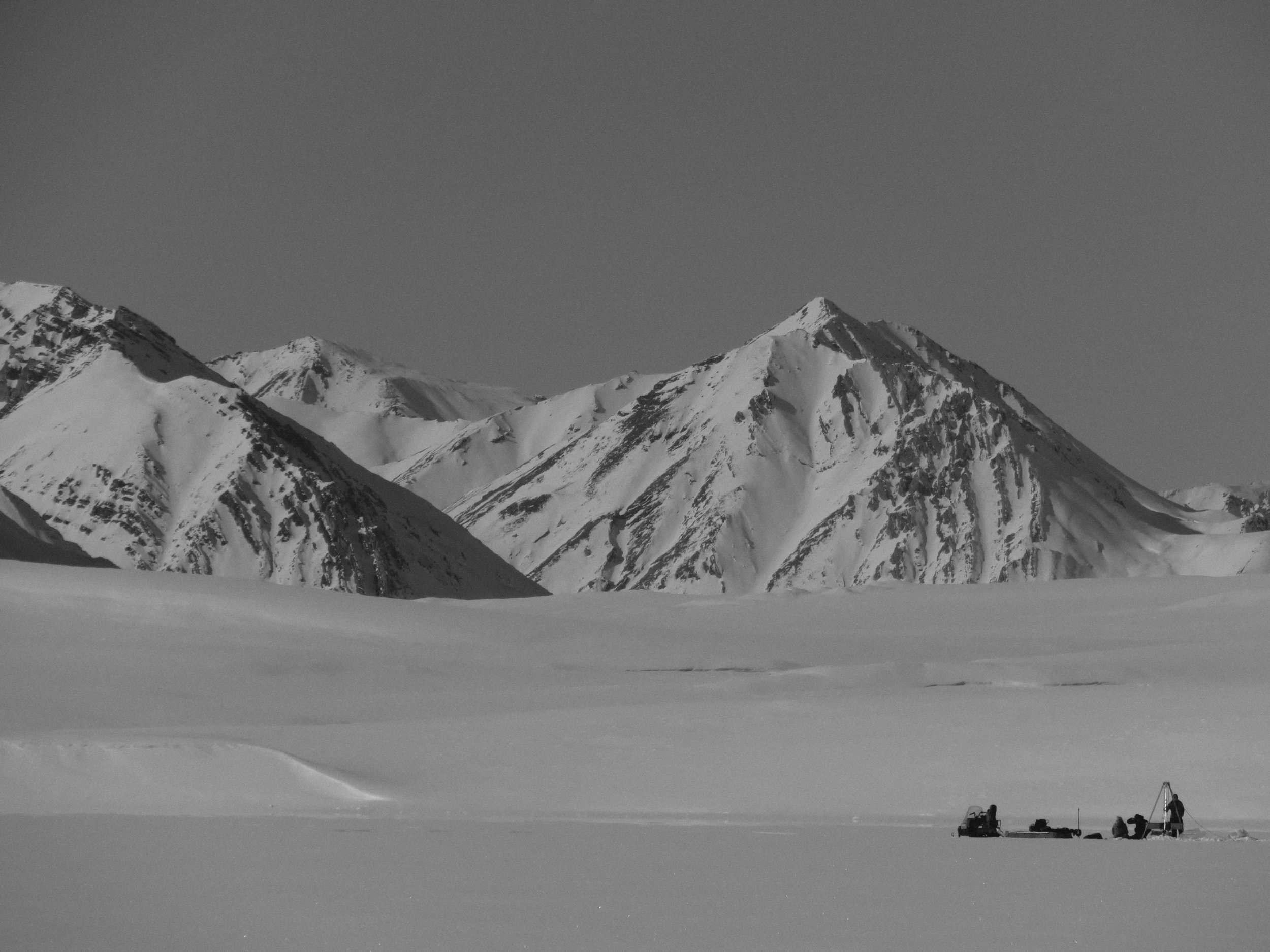 A snowy mountain landscape with a group of people snow camping on a flat ice field, with mountains in the background.
