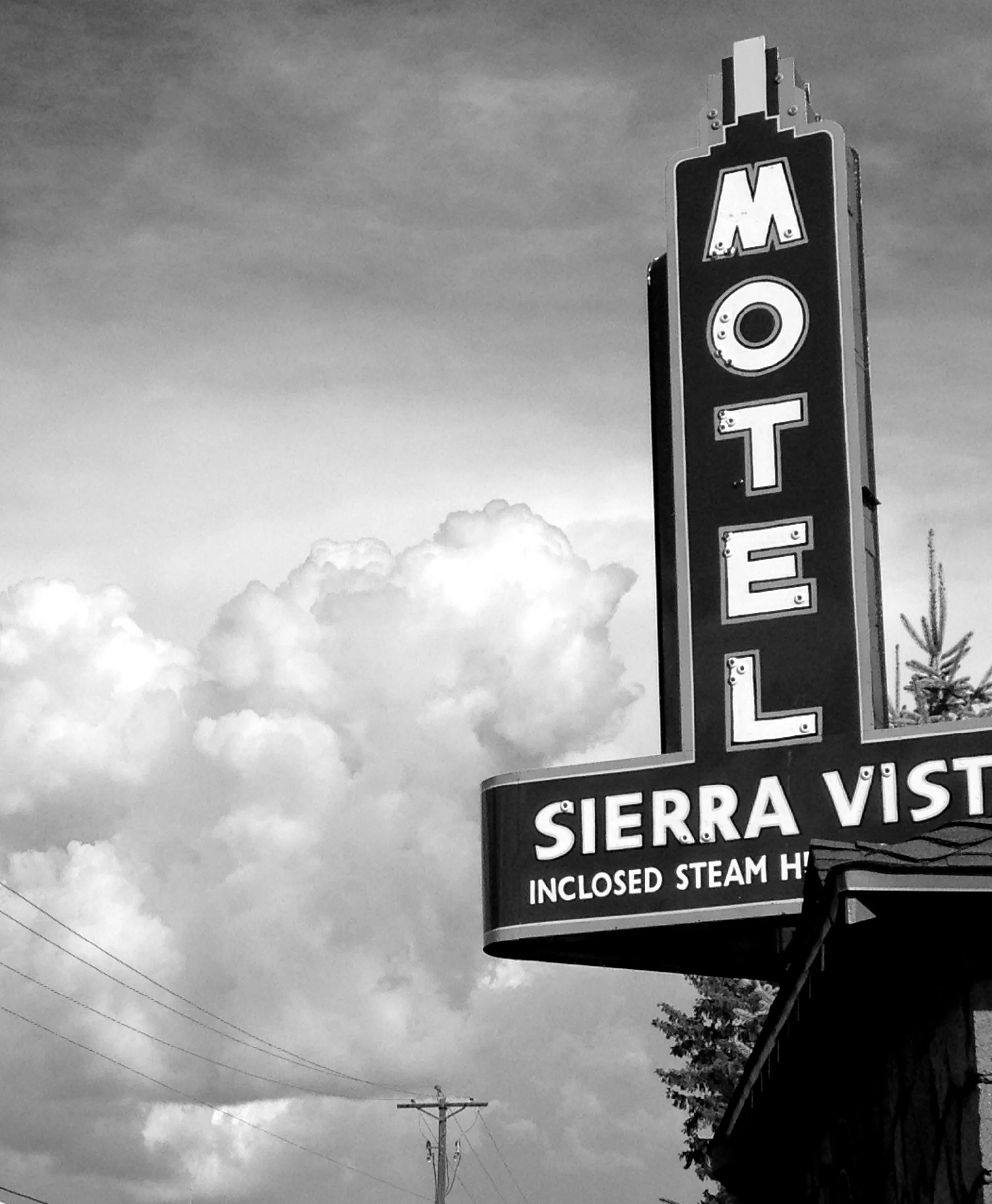Black and white photo of a vertical motel sign that reads 'MOTEL' and a horizontal sign below that says 'SIERRA VISTA INCLOSED STEAM H'. The sky in the background has large clouds and there are utility poles with wires.