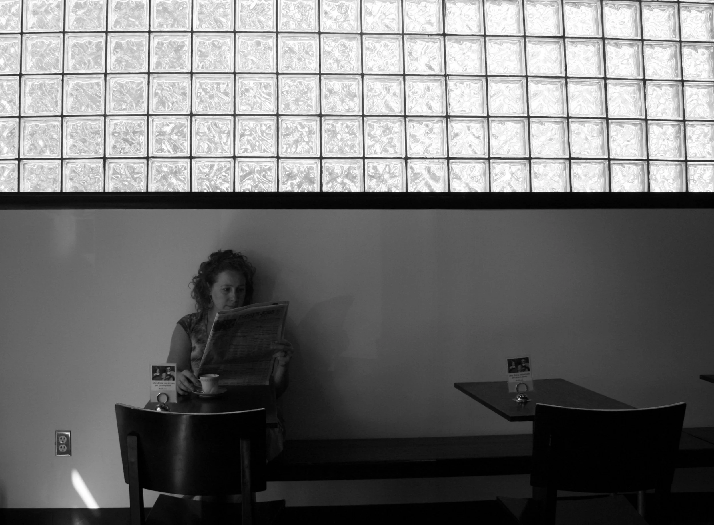 A woman with curly hair sitting alone at a table in a cafe, reading a newspaper and drinking coffee, with empty chairs nearby, in a black and white photo with a glass block window above her.