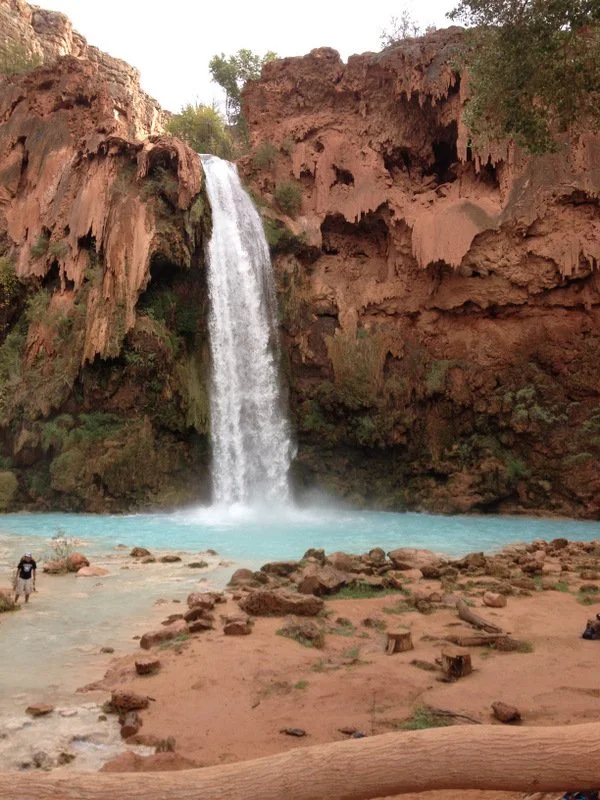 A tall waterfall cascading into a turquoise pool surrounded by rocky terrain and rugged red canyon walls with sparse vegetation.