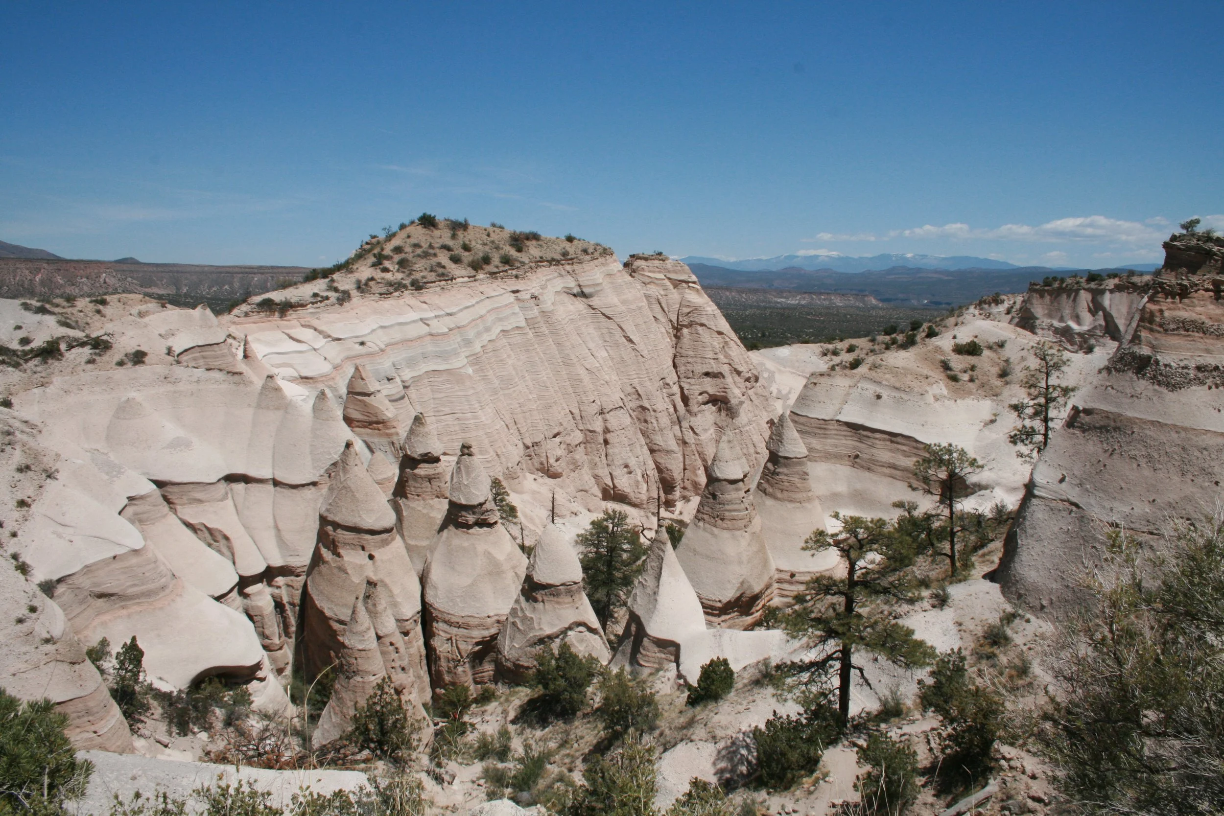 Barren rocky desert landscape with hoodoo rock formations, sparse trees, and distant mountains under a clear blue sky.