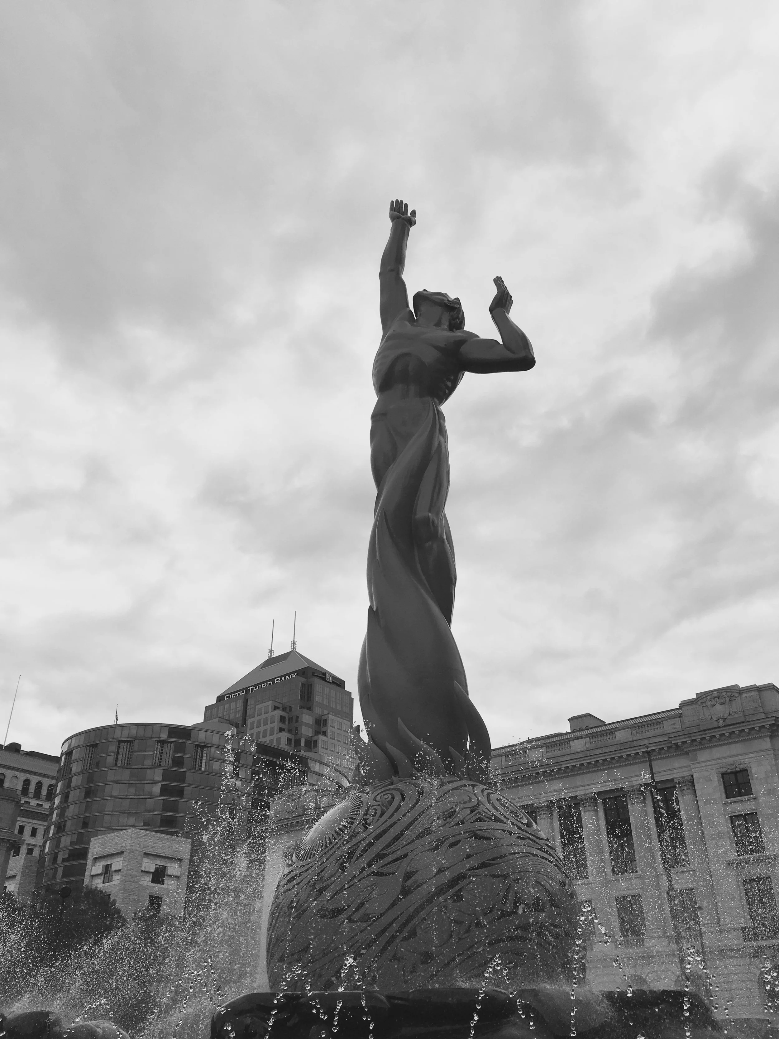 Black and white photo of a statue of a woman with arms raised, standing on a globe, surrounded by water in a fountain.