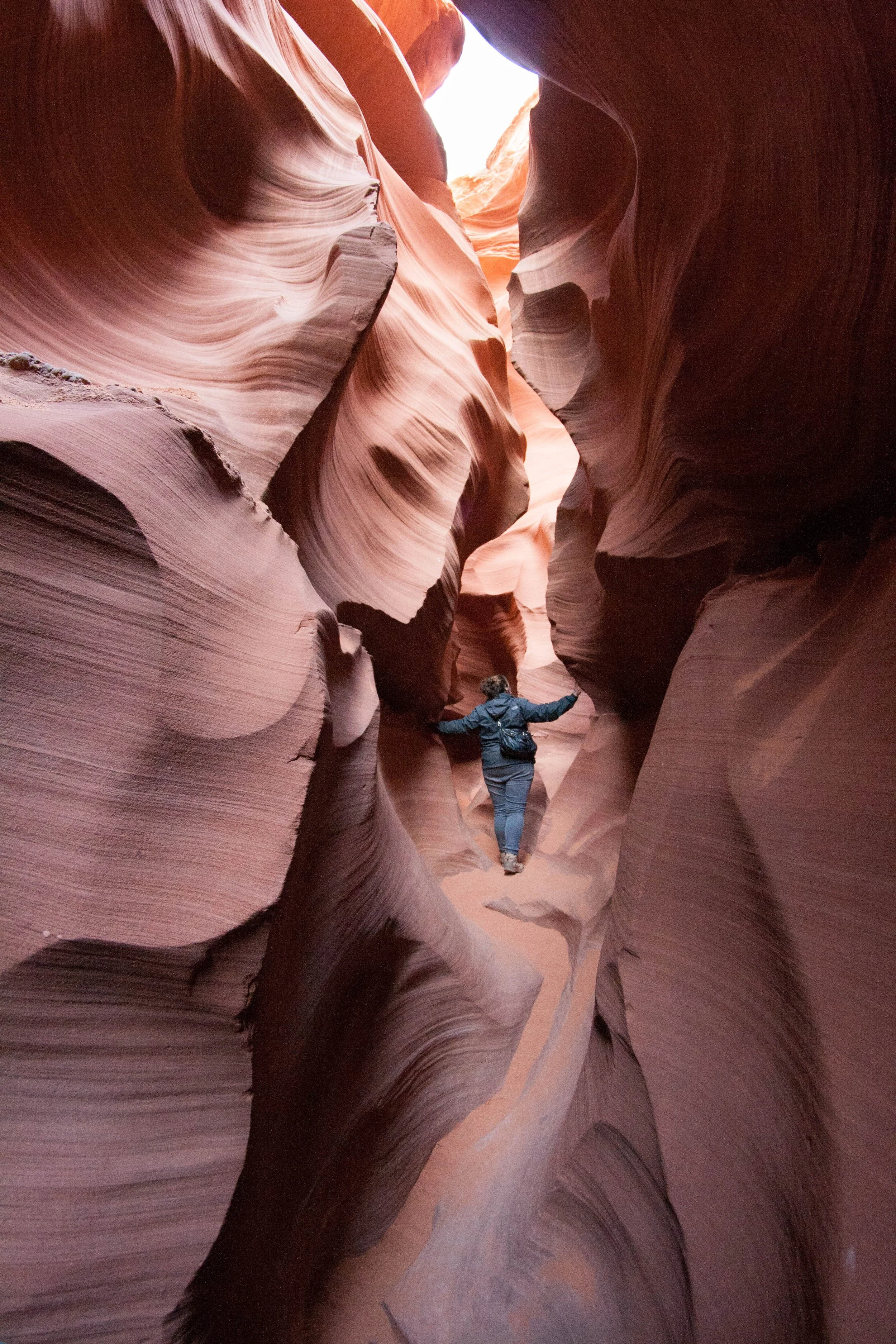 Person walking through narrow slot canyon with smooth, layered rock walls in shades of pink, orange, and brown.