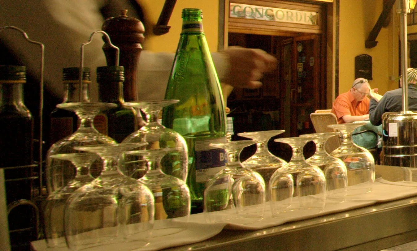 Close-up of upside-down wine glasses and a green glass bottle on a table inside a restaurant with diners in the background.