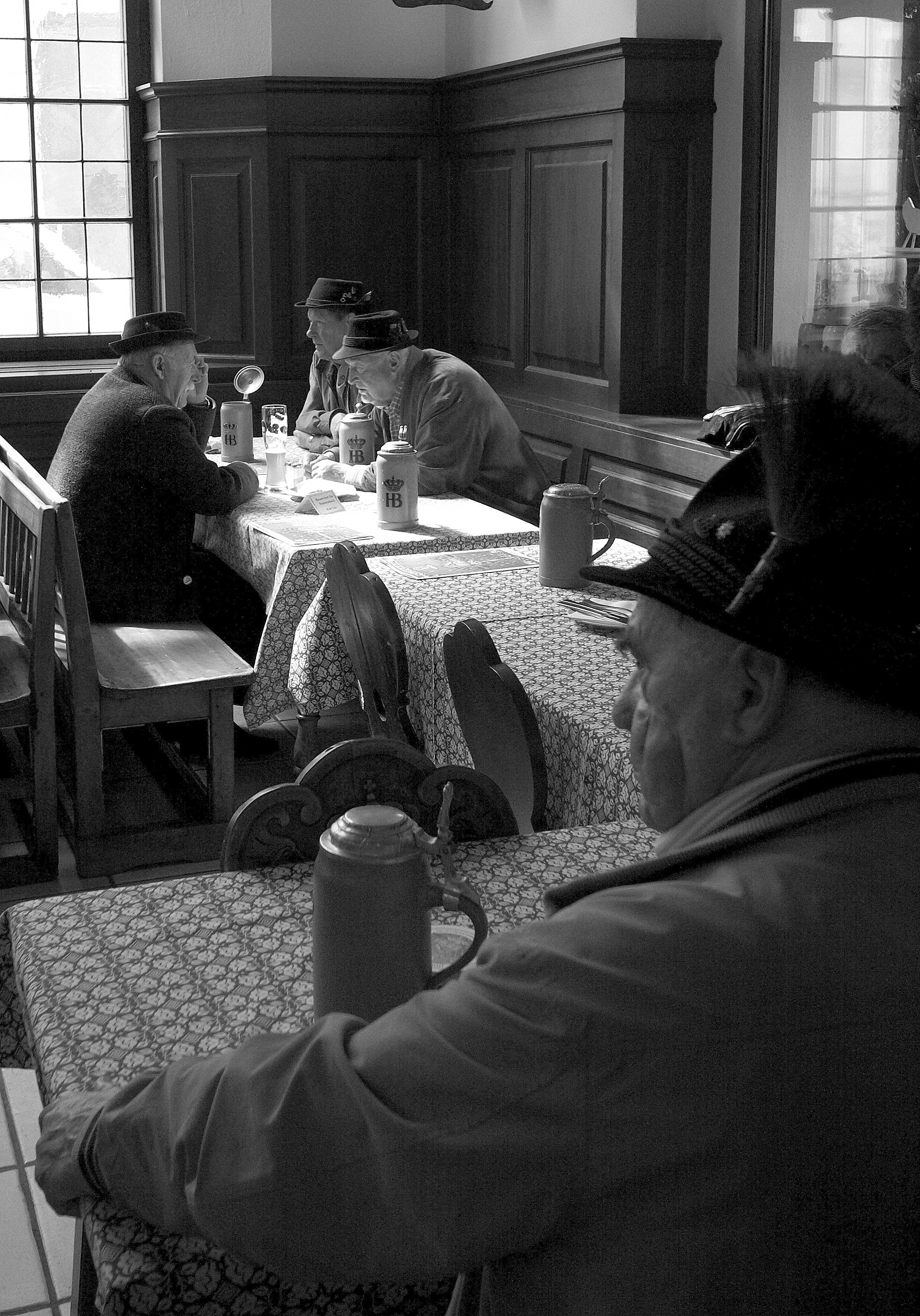 Four elderly men wearing hats sitting at a table in a restaurant, engaged in conversation, with traditional beer steins on the table, and a large window providing natural light.