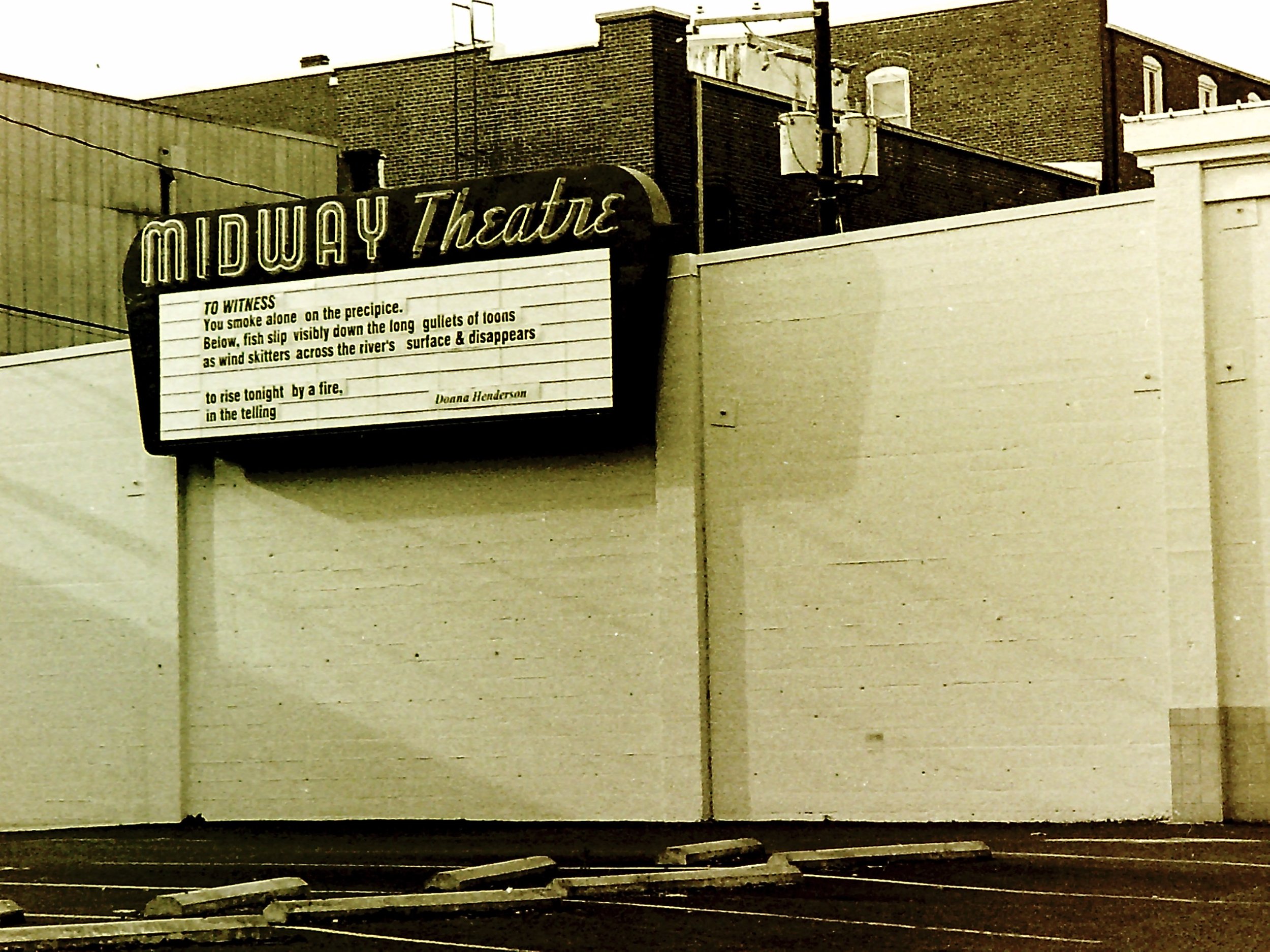 An empty parking lot in front of a building with a movie theater marquee for the Midway Theatre, displaying a poetic message about witnessing a fire.