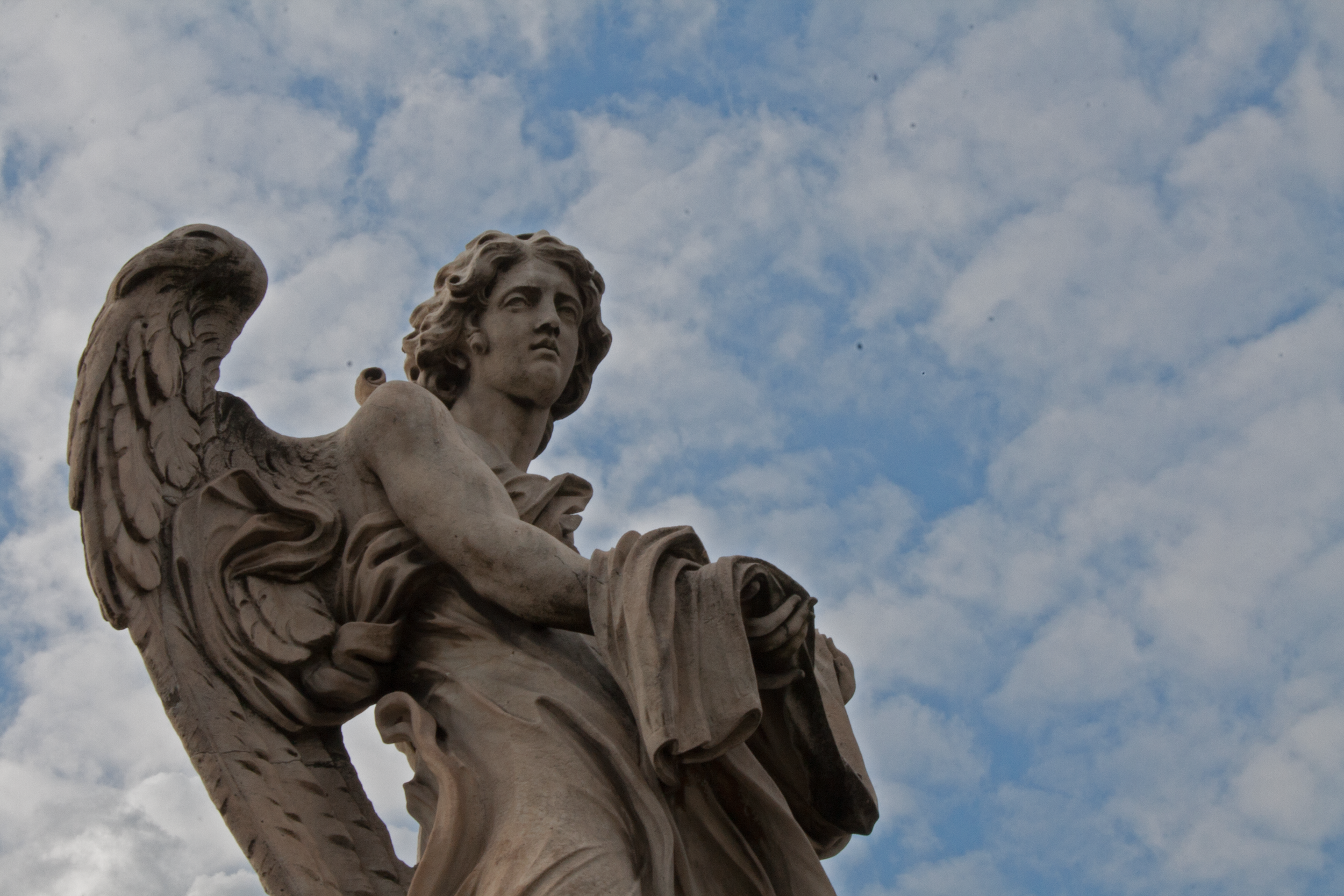 A stone sculpture of an angel with detailed wings, long curly hair, and a serious expression, set against a partly cloudy sky.