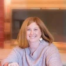 A woman with red hair and a gray top smiling in a room with a television and wooden furniture.
