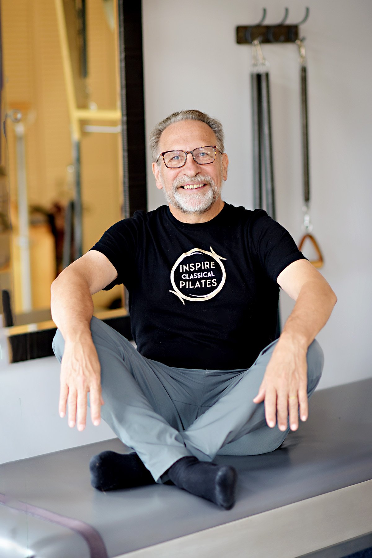 Smiling older man with glasses, grey hair, and beard, sitting cross-legged on a Pilates reformer machine in a fitness studio, wearing a black Inspire Classical Pilates t-shirt.