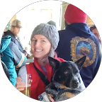 A woman smiling and wearing a gray beanie, holding a black dog, surrounded by people dressed warmly in outdoor winter clothing.