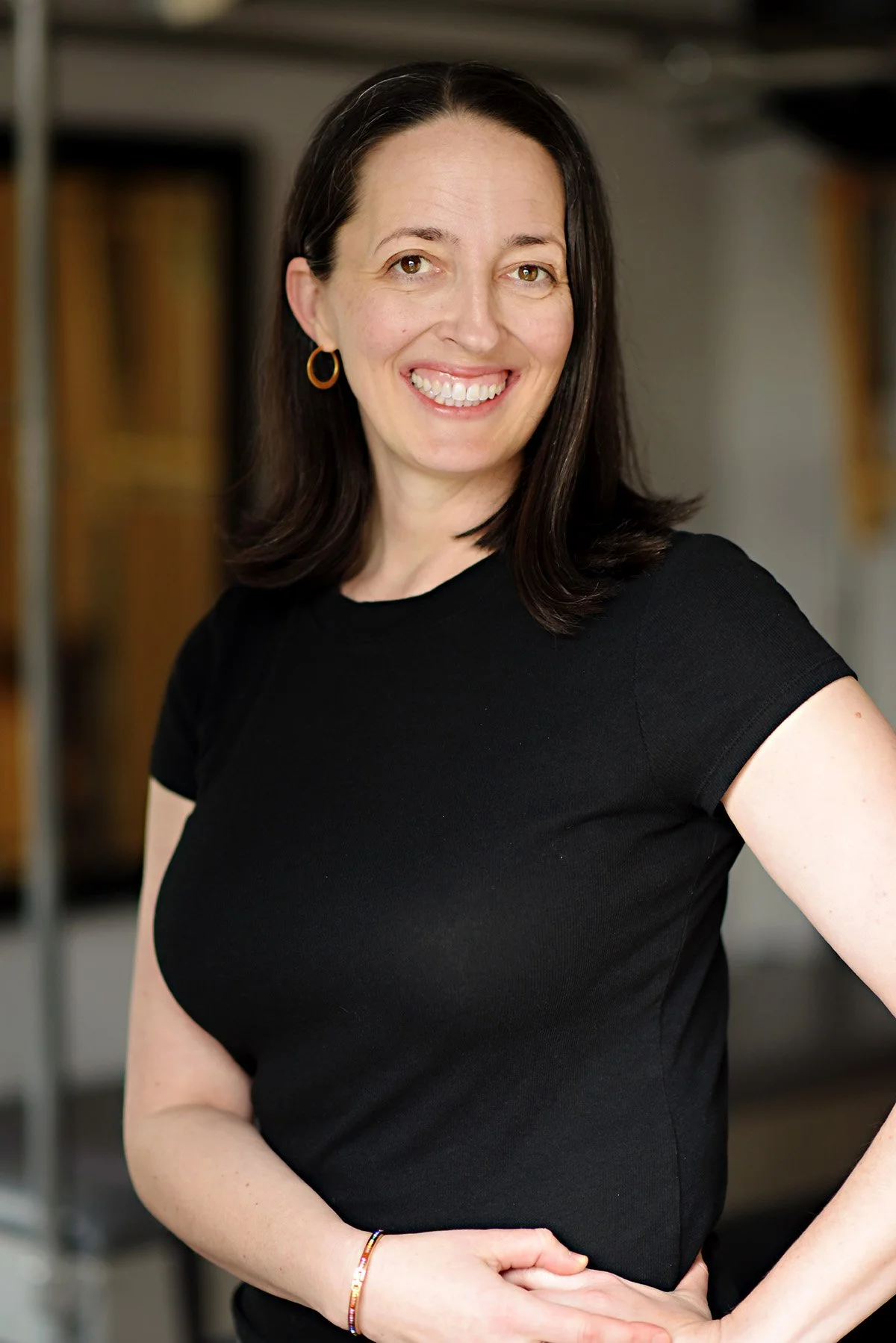 A woman with medium-length dark hair, wearing a black shirt and gold hoop earrings, smiling and standing with her hand on her hip in an indoor setting with blurred bookshelves in the background.