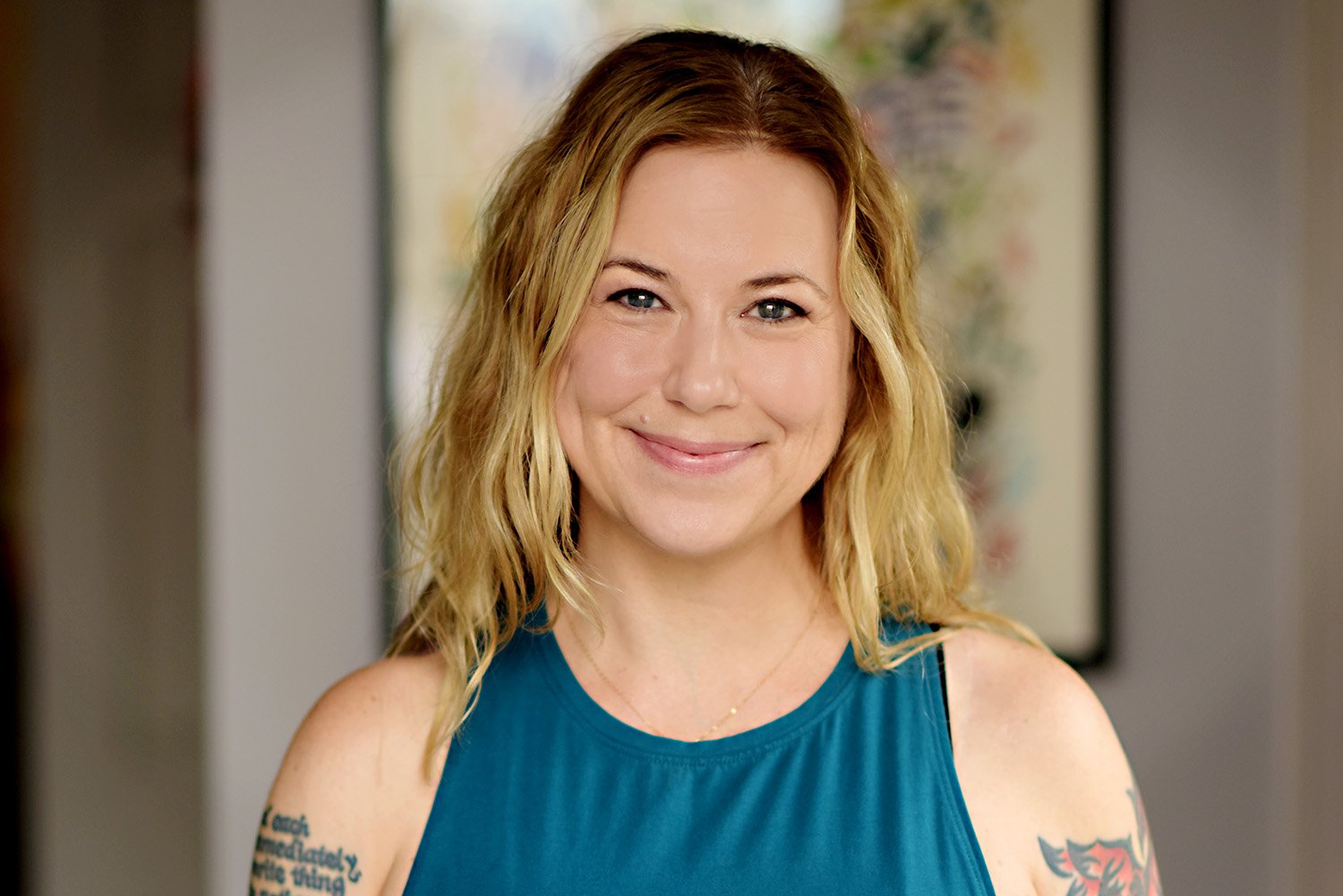 Woman with blonde hair smiling in a blue sleeveless top, standing in front of a colorful framed artwork in a neutral-colored room.