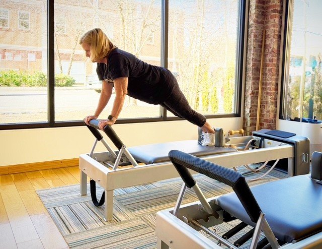 A dimly lit Pilates reformer machine on a wooden floor in an empty room with large windows and exposed brick walls.