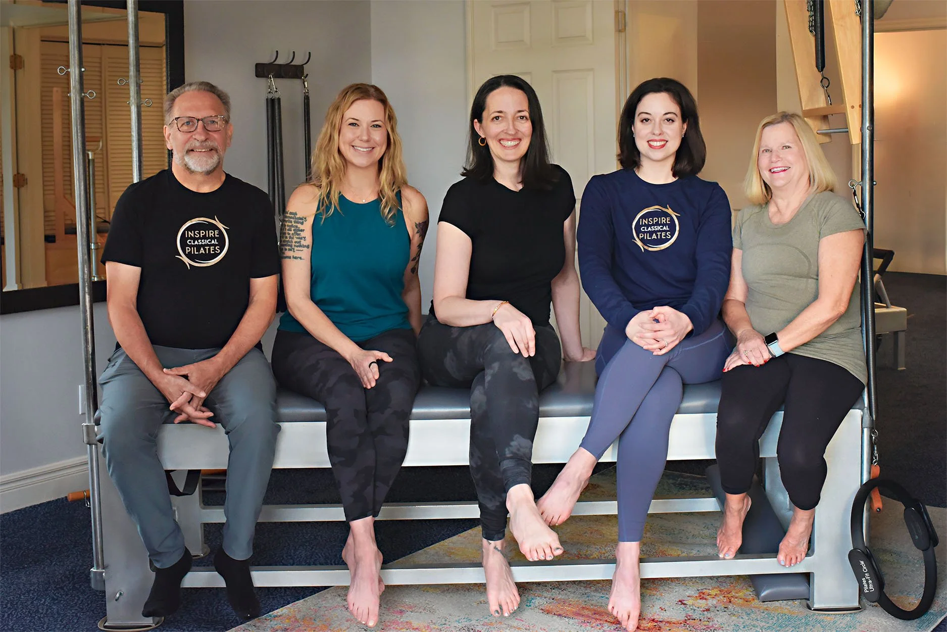 Five people sitting on a reformer machine in a Pilates studio, smiling at the camera. The first person on the left is an older man wearing glasses and a black T-shirt with the logo 'Inspire Classical Pilates.' The second person is a woman with blonde hair wearing a teal sleeveless top. The third person is a woman with dark hair wearing a black T-shirt. The fourth person is a woman with dark hair wearing a navy blue 'Inspire Classical Pilates' sweatshirt. The fifth person is an older woman with blonde hair wearing a green T-shirt and black leggings, with a smartwatch on her wrist.
