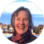 Smiling woman with short gray hair, wearing a purple and black scarf, outdoors near water with boats and buildings in the background.