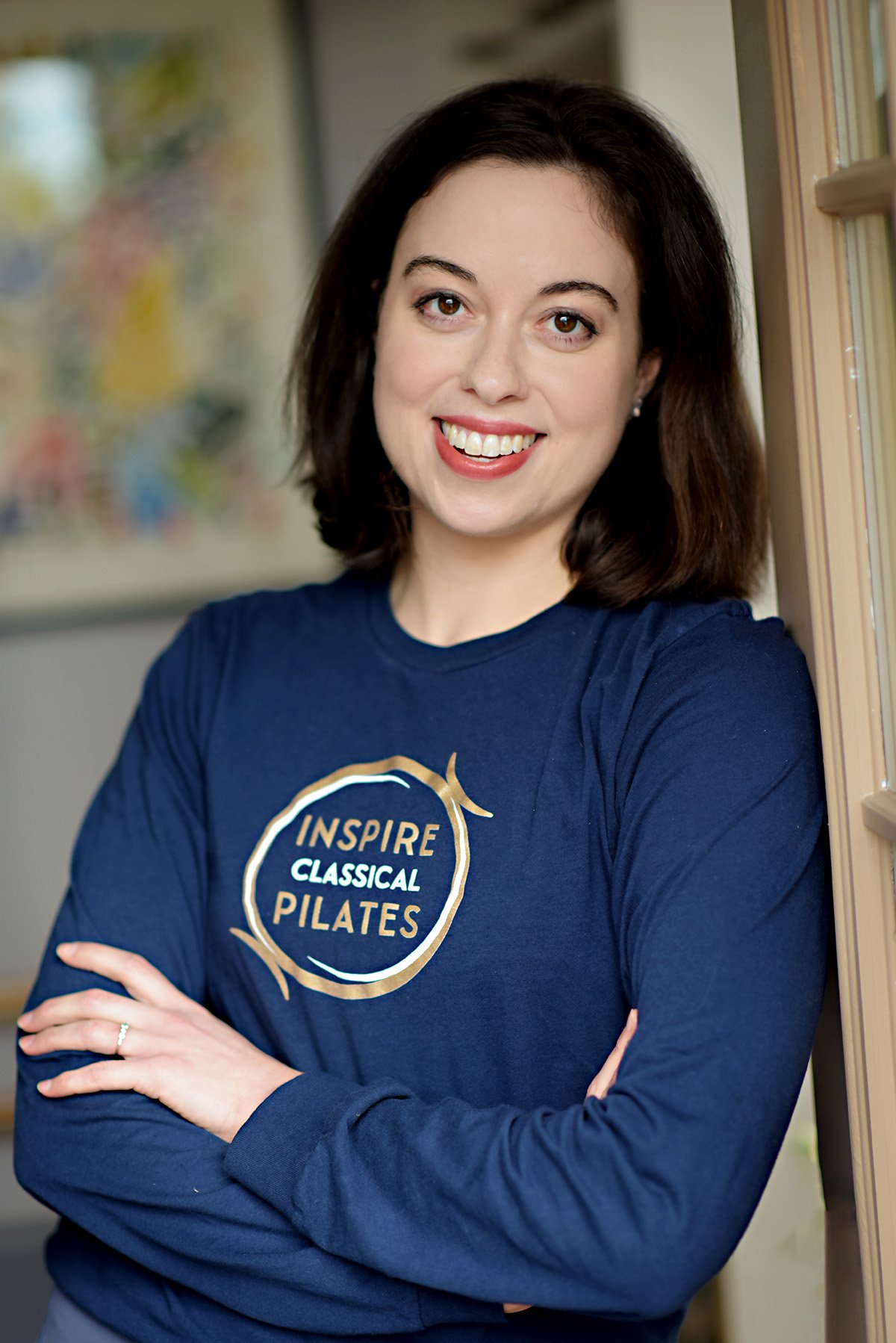 A young woman with shoulder-length dark hair, smiling and wearing a navy blue long-sleeve shirt that says "INSPIRE CLASSICAL PILATES" in a circular logo.