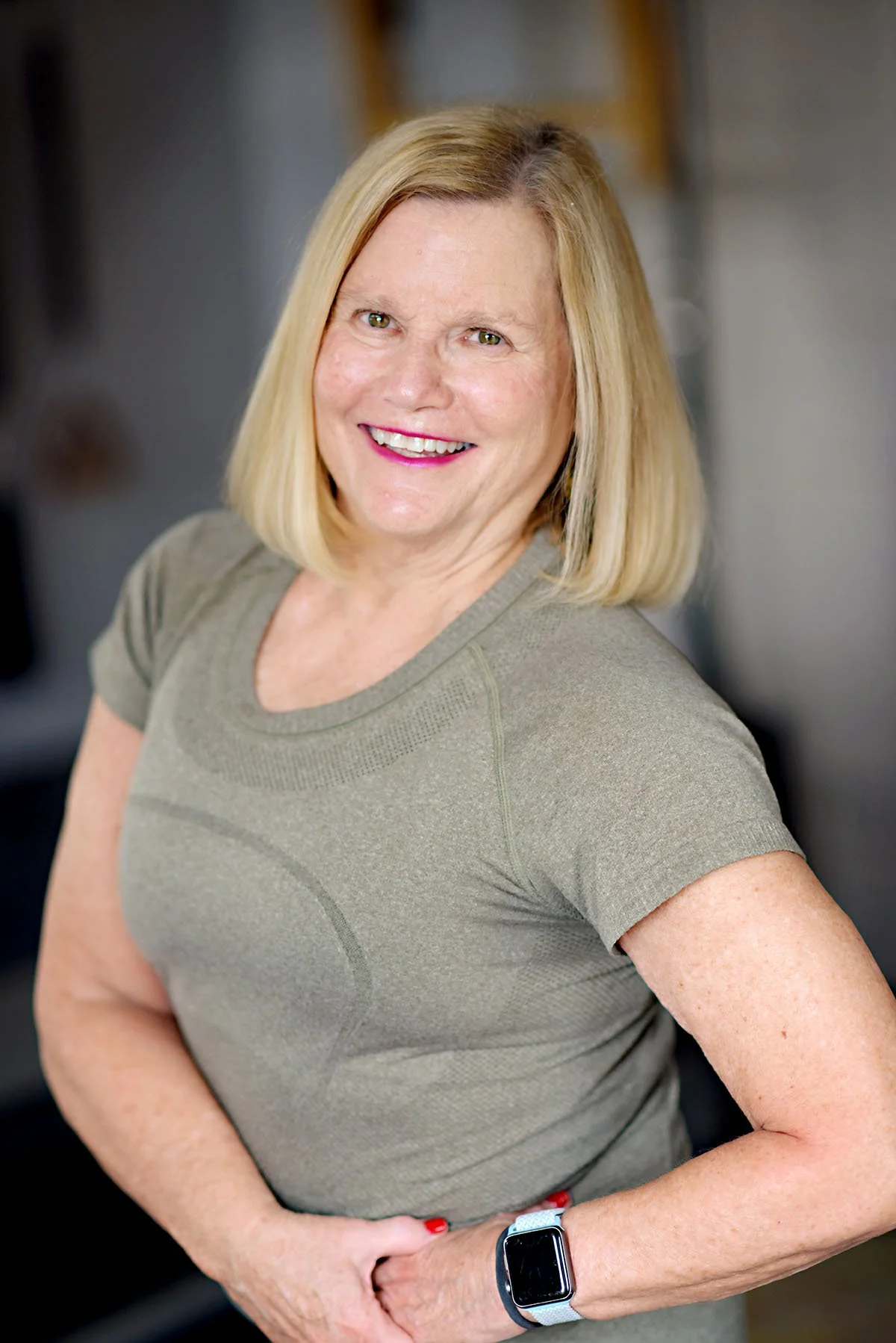A smiling middle-aged woman with blonde hair, wearing a short-sleeved olive-green athletic shirt and a smartwatch, standing indoors with a blurred background.