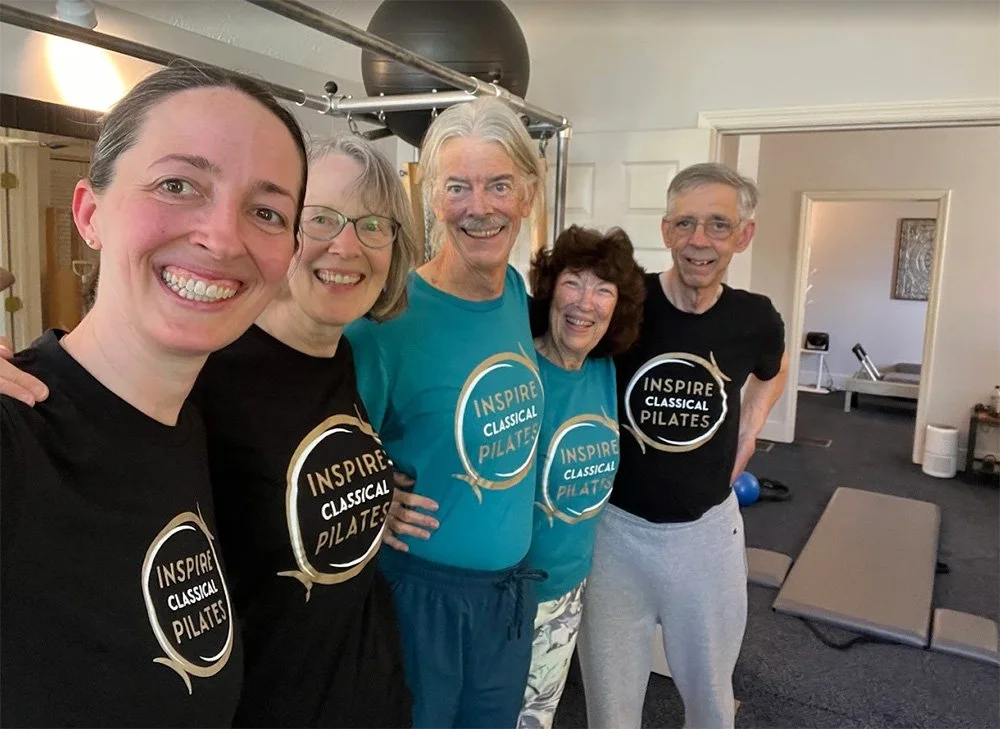 Five people smiling and posing together in a fitness or pilates studio, wearing matching Inspire Classical Pilates shirts.