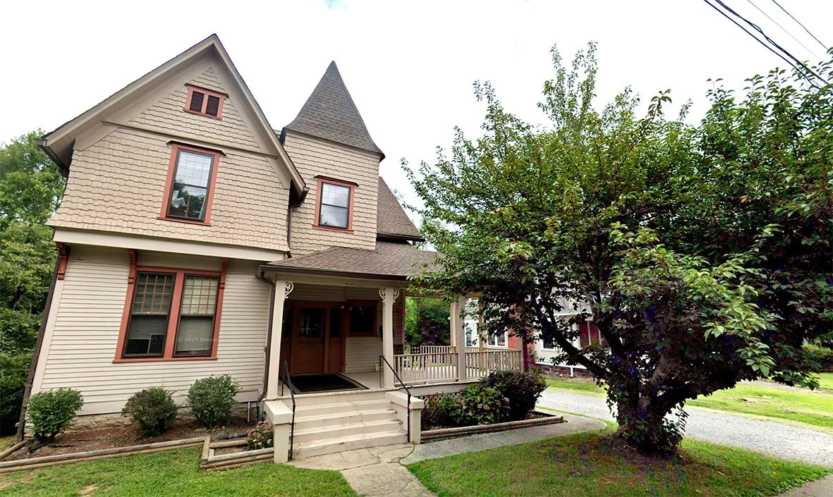 A Victorian-style house with a tower, front porch, and trimmed bushes in a residential neighborhood.