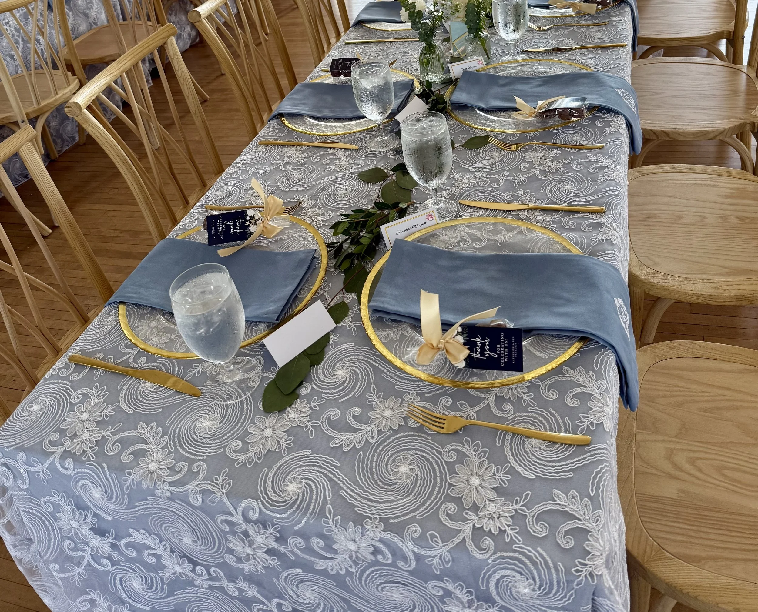 Elegant dining table set for a formal event with blue napkins on gold-rimmed plates, gold-colored cutlery, water glasses, and green leaf decorations on a white embroidered tablecloth rented in Omaha, NE.