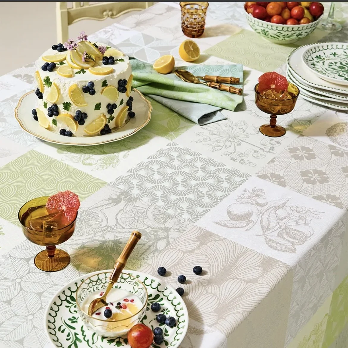 A table set for a meal with a lemon cake decorated with blueberries and lemon slices in the center, surrounded by bowls of fruit, plates, glasses, and a bowl of cherries. There are colorful napkins and a pastel patterned tablecloth.