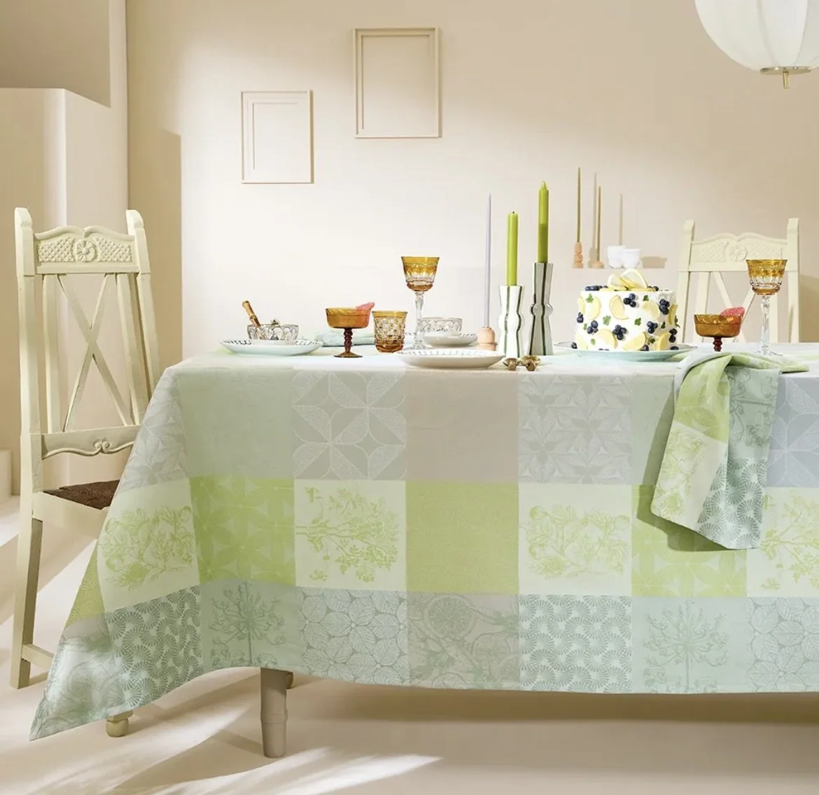 A dining table set for a celebration with a white cake on a pastel green, blue, and yellow high-end linen for rent in Omaha, NE from Akita Event Rentals and amber-colored glassware, in a bright, minimally decorated room.