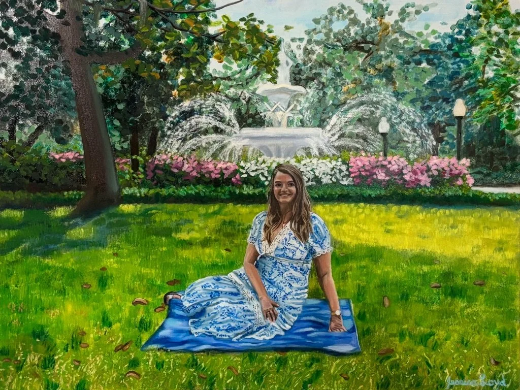 A young woman in a blue and white dress sitting on a blue mat on a grassy lawn in a park with a fountain and trees in the background.