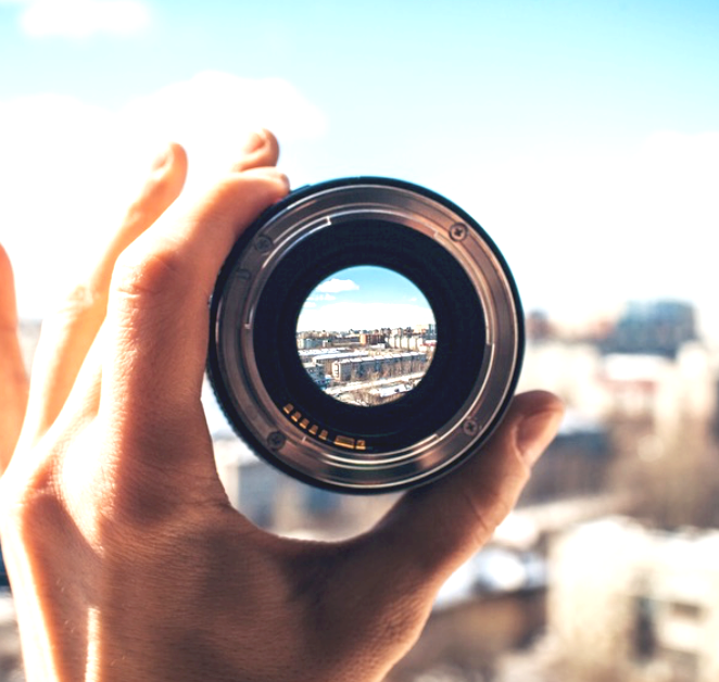 Person holding a camera lens facing outward with a cityscape in the background, viewed through the lens.